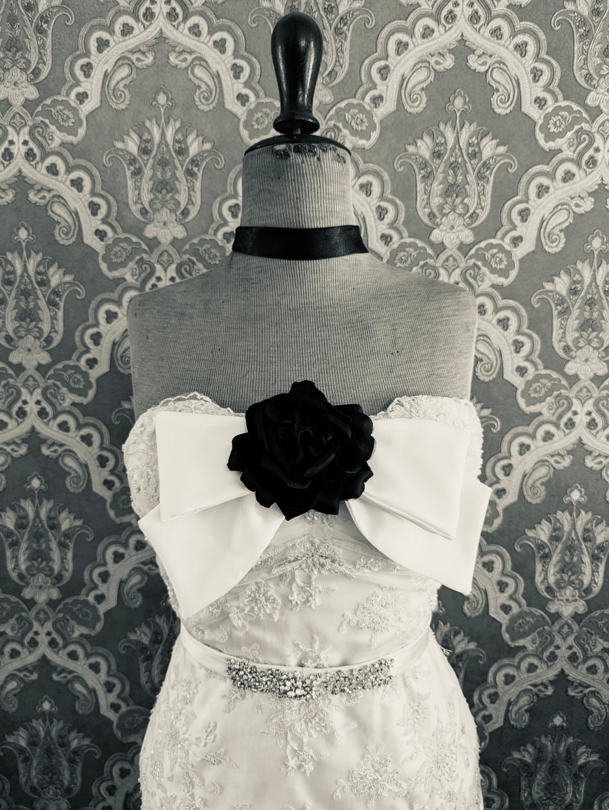 Broochella  dolce amore handmade bridal brow brooch in ivory silk satin. There is a black fabric flower embellishment at the centre of the bow. Pictured on a mannequin that is wearing a white, beaded strapless wedding dress. The background is green and gold damask wallpaper. 