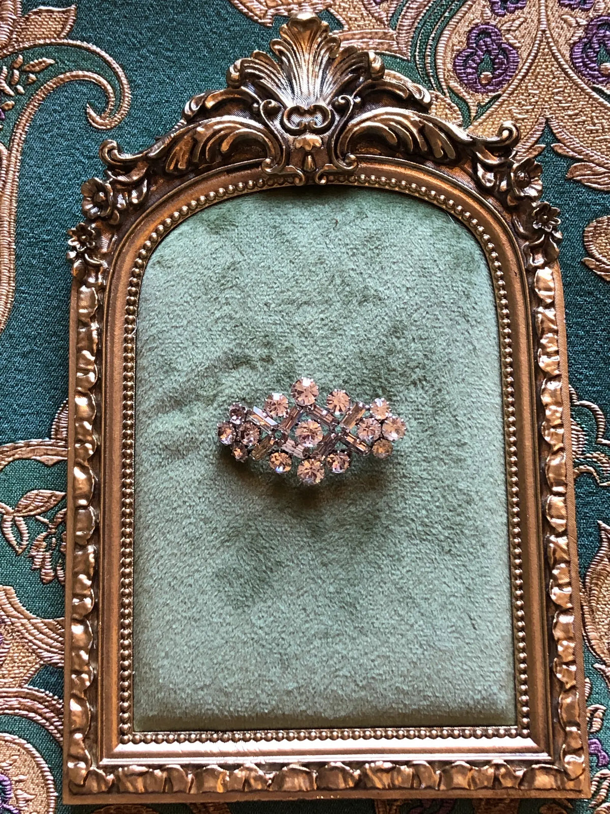 Broochella Donatella Vintage Crystal Lattice Brooch featuring round and baguette-cut clear crystals in an open geometric silver-tone setting. Pictured on a green velvet tray with an ornate gold frame. The background is green and gold damask wallpaper