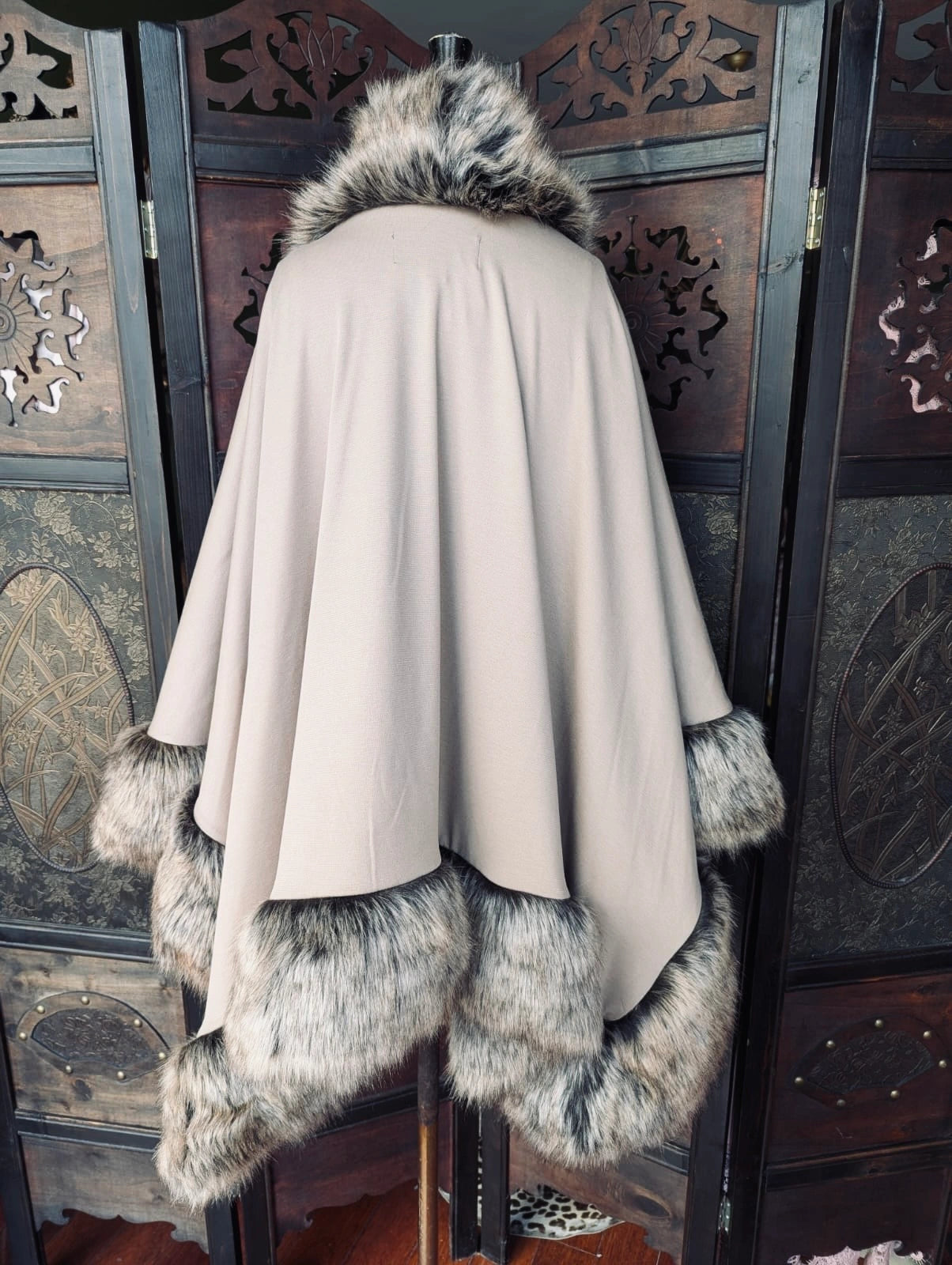 A Broochella Stylist's Closet Donna Salyers Fabulous Furs OSFA beige cape with a large dark brown and gray faux  fur collar, cuffs, and hem, displayed on a mannequin in front of a dark wooden decorative screen.