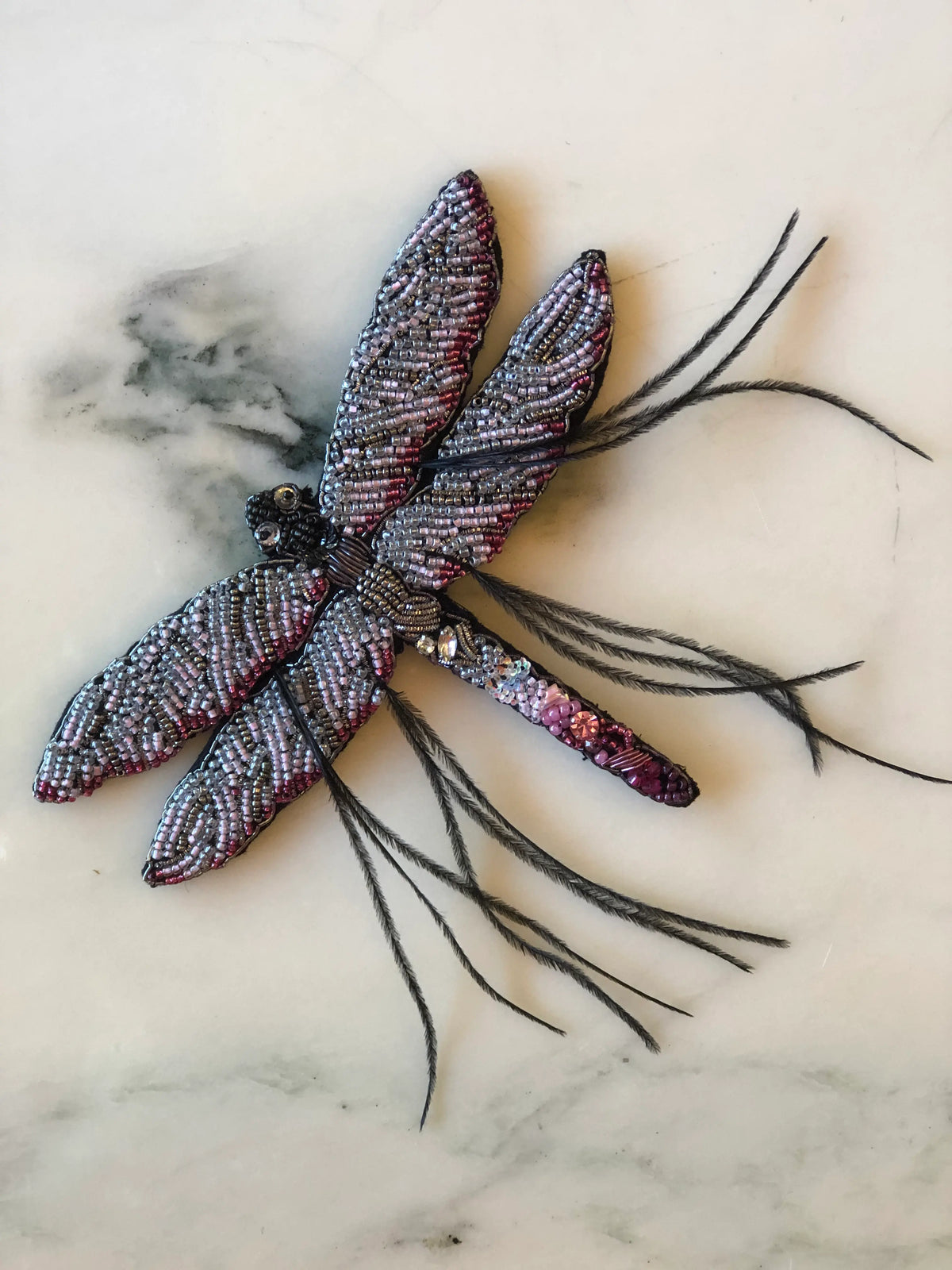 Broochella Dragonfly Bridal Brooch featuring intricate silver, blush pink and metallic beadwork with sparkling crystal details and delicate black feather accents, photographed on a pale cream marble background.