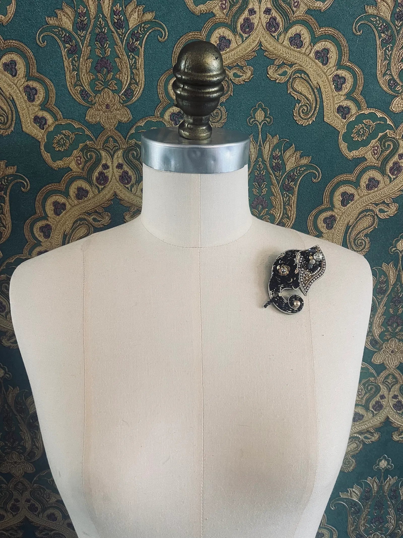 Broochella hand beaded black and gold elephant brooch. Pictured on a mannequin. The background is green and gold damask wallpaper