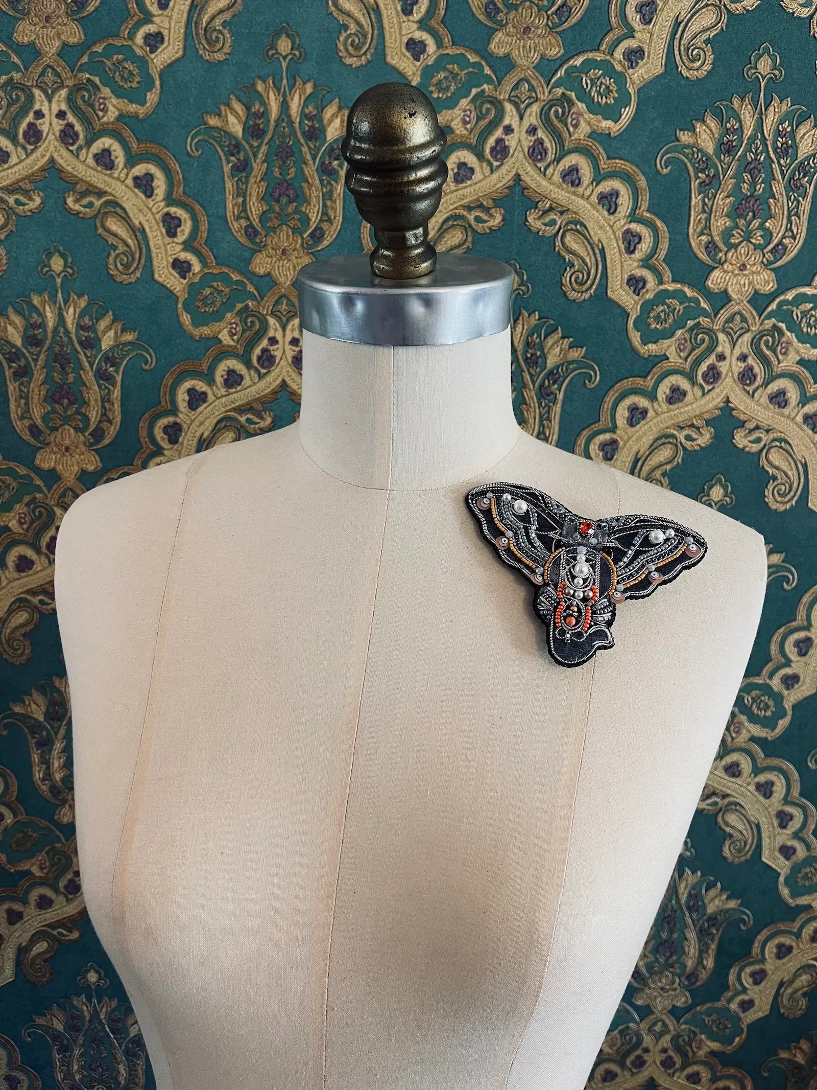Broochella elephant face brooch hand made from  beads and sequins. Pictured on a mannequin. The background is green and gold damask wallpaper.