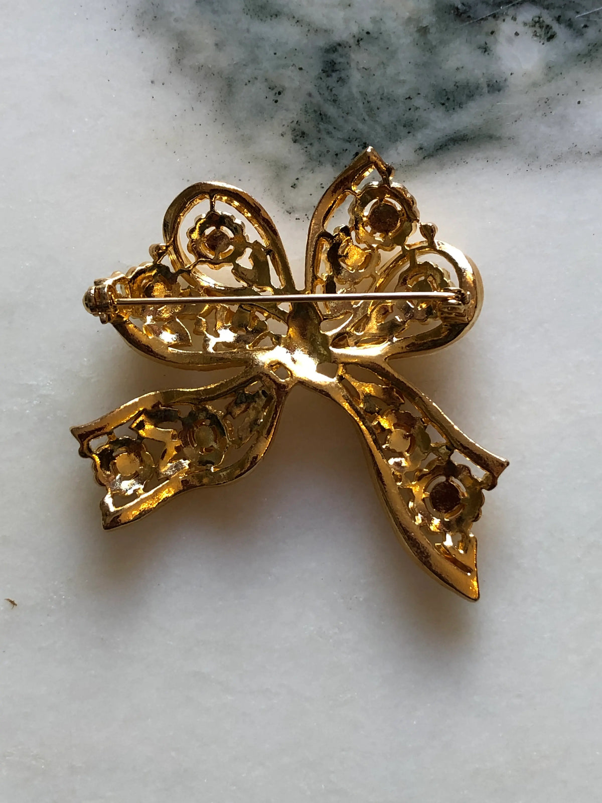 Broochella Elisabetta Vintage Gold Bow Brooch featuring an ornate sculptural ribbon design with floral and scroll openwork detailing on a cream and grey marble background.