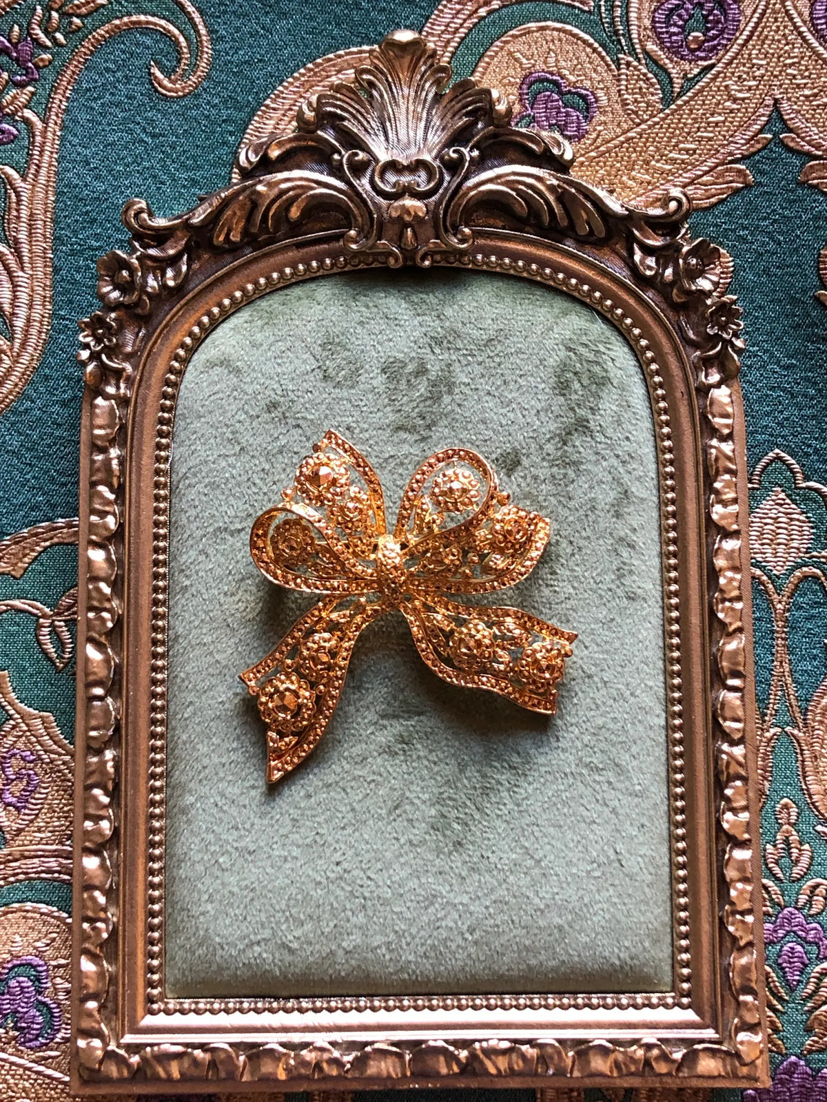 Broochella Elisabetta Vintage Gold Bow Brooch featuring an ornate sculptural ribbon design with floral and scroll openwork detailing. Pictured on a green velvet tray with an ornate gold frame. The background is green and gold damask wallpaper. 