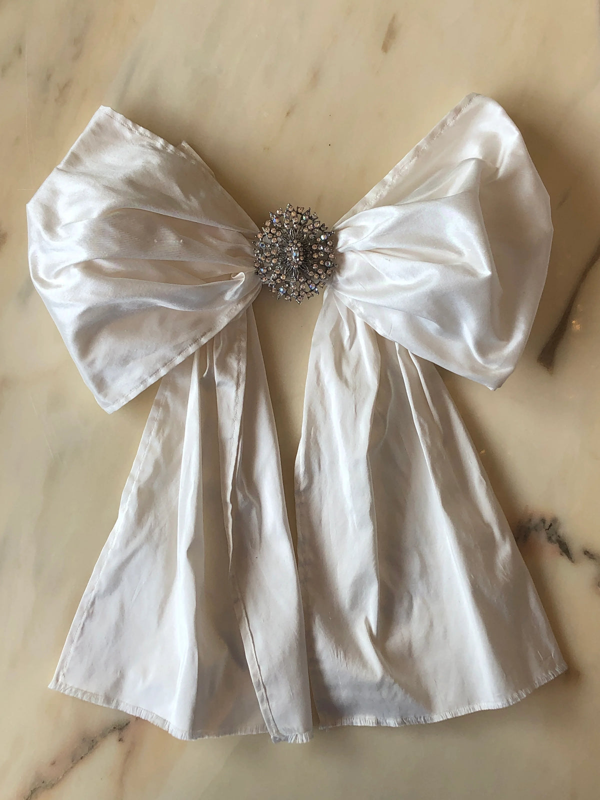 Broochella Embellished Ivory Bridal Bow featuring an oversized ivory satin bow with long flowing tails and an ornate crystal brooch centre, photographed on a pale cream marble background.