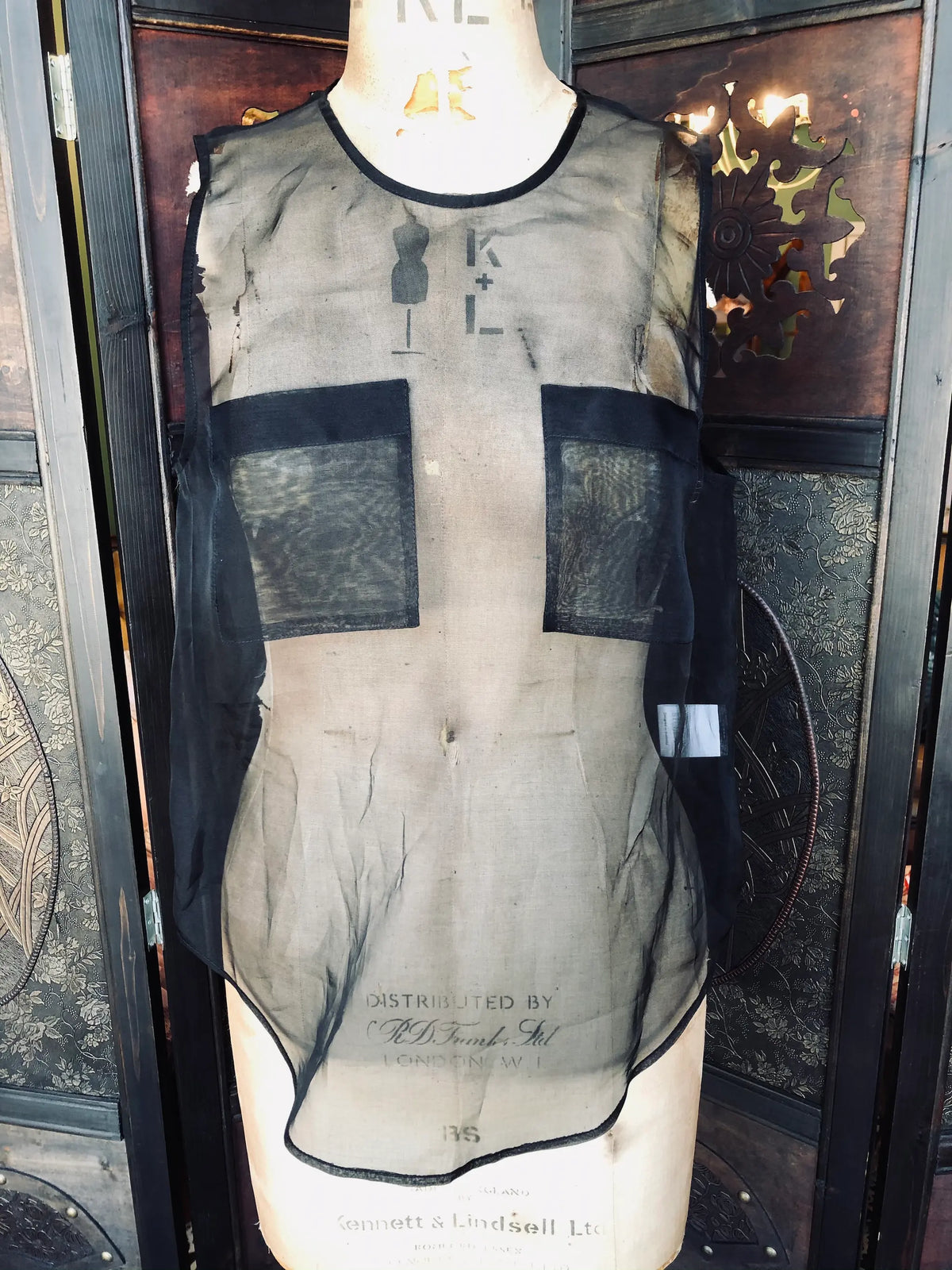 Broochella Emerson studios, Small, 100% Silk, Black, Sleeveless design, Sheer silk finish, Front panel pockets. Keyhole back detail. It is presented on a vintage mannequin. The background is a ornate, vintage, dark wood room divider. 