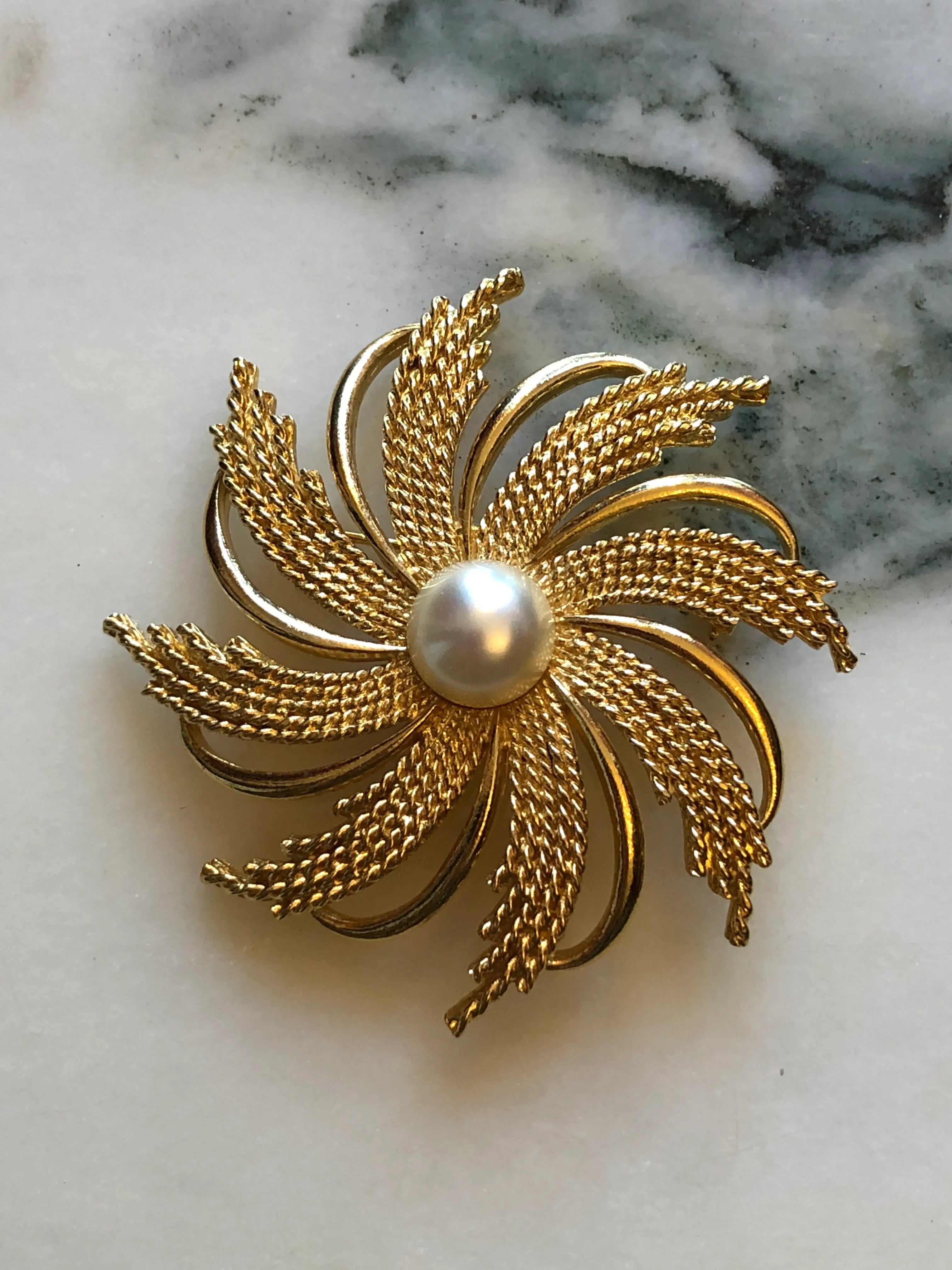 Broochella Emma Page vintage sunburst gold-tone brooch with woven mesh rays, sculptural loops and a faux pearl centre. The background is white marble