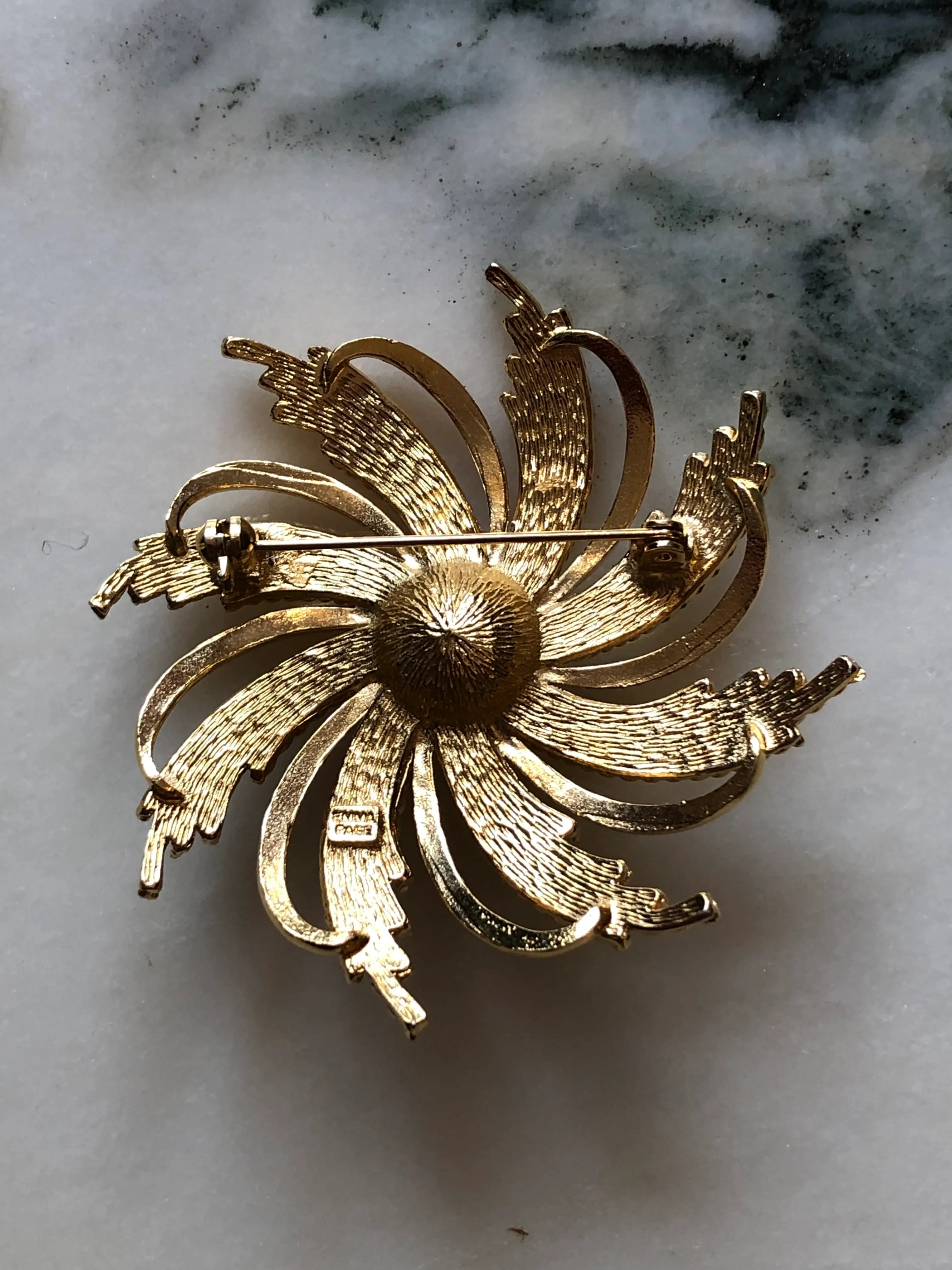 the back view of Broochella Emma Page vintage sunburst gold-tone brooch with woven mesh rays, sculptural loops and a faux pearl centre. The background is white marble