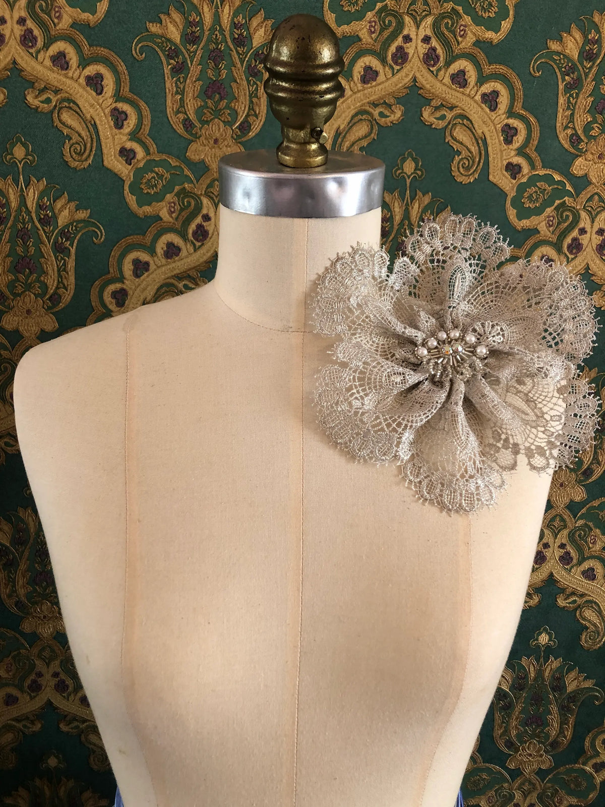 Broochella Étoile de Dentelle handmade designer silver-grey lace flower brooch styled at the shoulder of a cream dressmaker’s mannequin, featuring layered lace petals, pearl accents and a crystal centre, against an ornate dark green and gold patterned textile background.