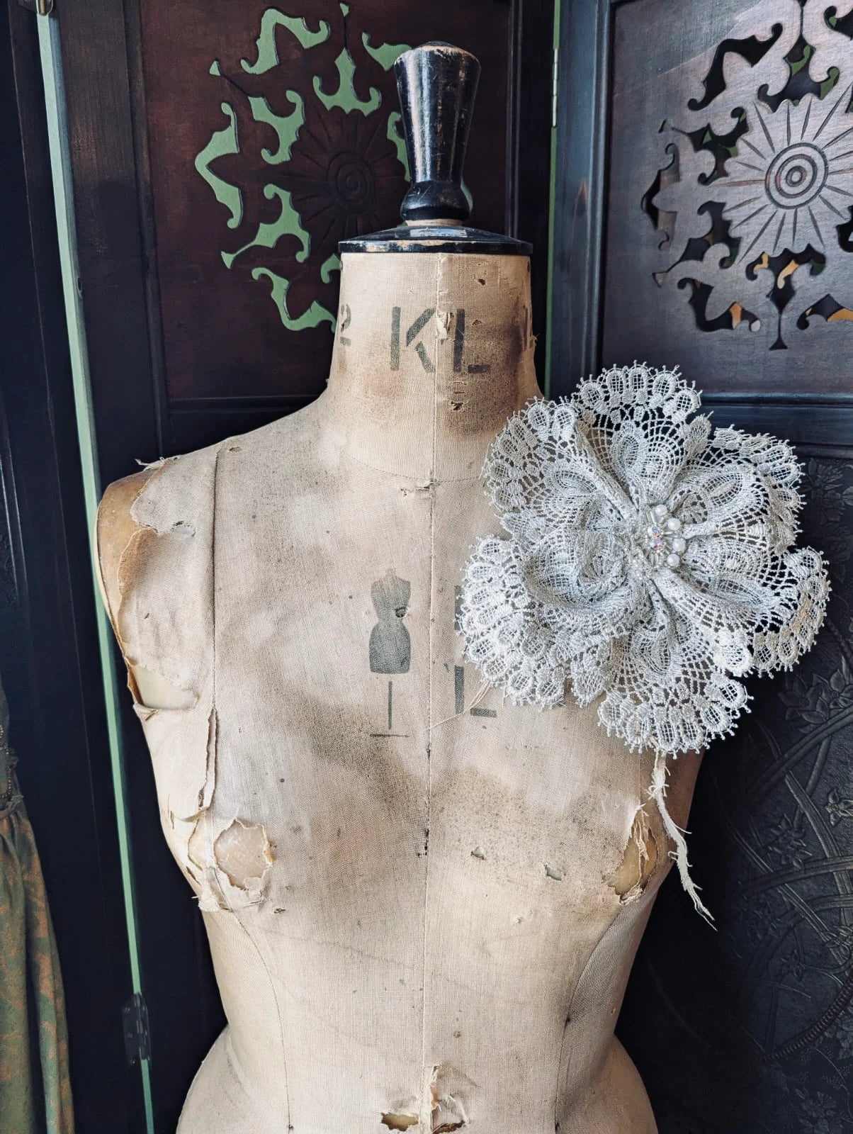 Broochella etoile de dentelle ivory lace handamde fabric flower brooch with beaded centre. It is pinned to an old mannequin. The background is a decorative brown wooden screen.