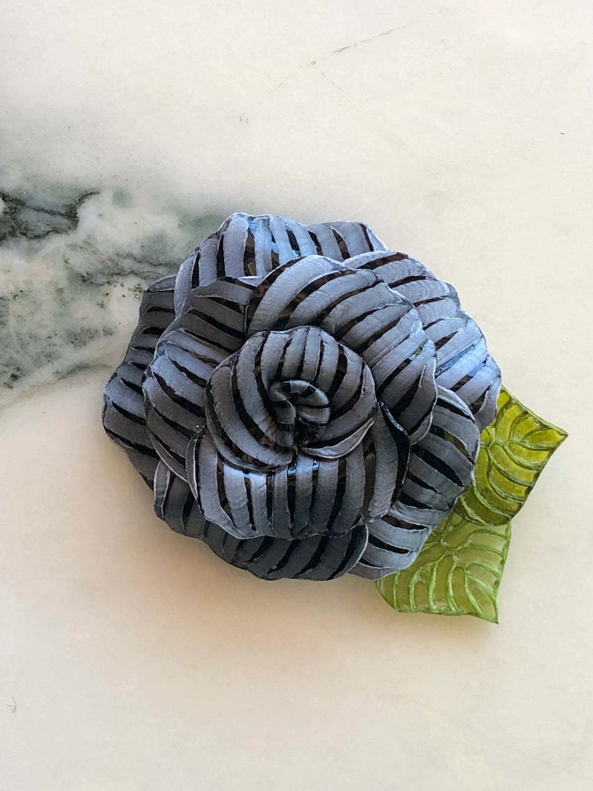 Broochella Fabrice Paris Camelia flower brooch for men in black and soft grey striped fabric, featuring layered sculptural petals and two textured green leaves. A distinctive men's floral lapel accessory photographed on white marble with grey-green veining.