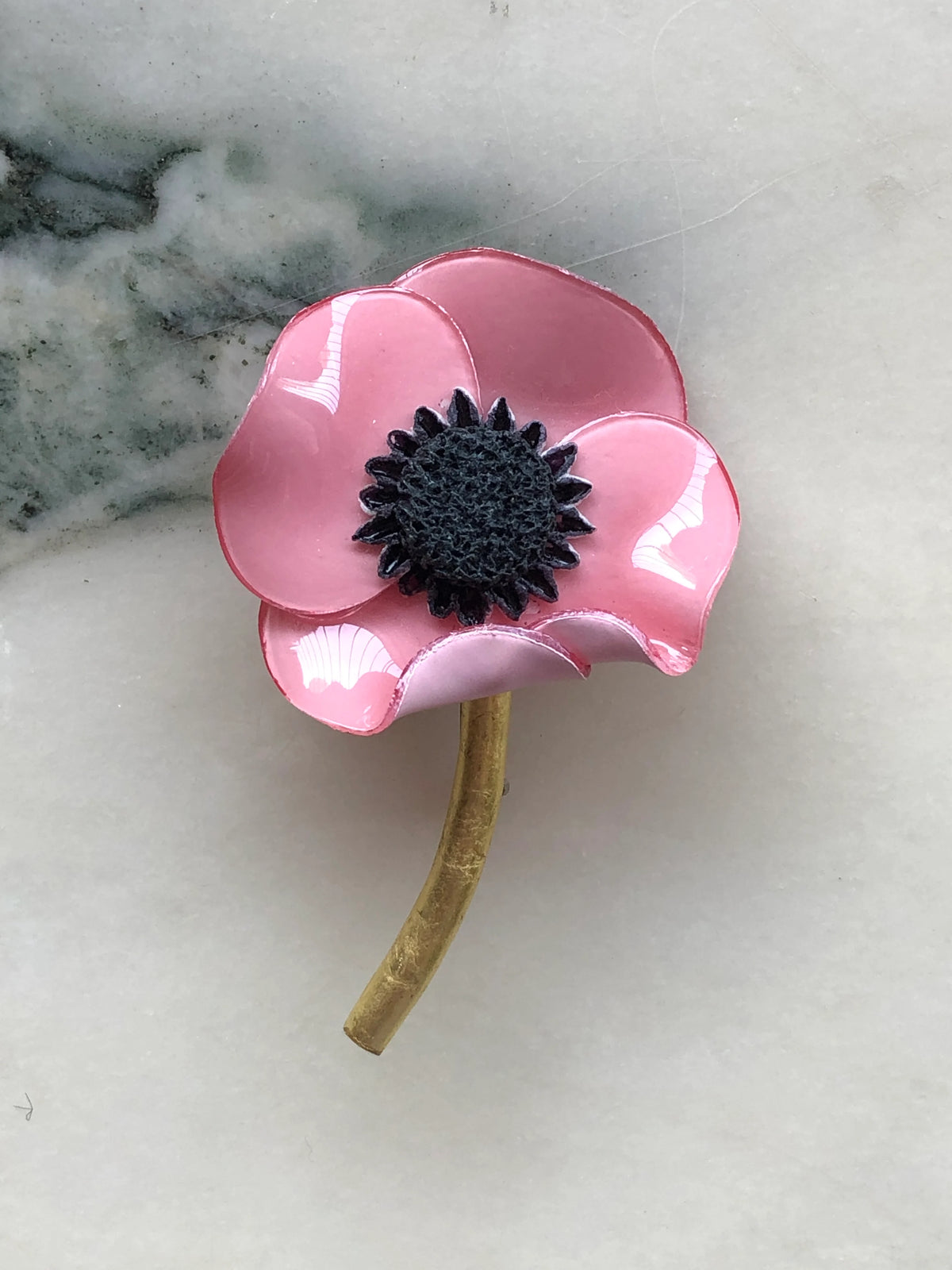 Broochella Fabrice Paris Poppy Flower Brooch in soft pink with glossy sculptural petals, a textured black centre and curved antique gold stem. Photographed on white marble with grey-green veining.