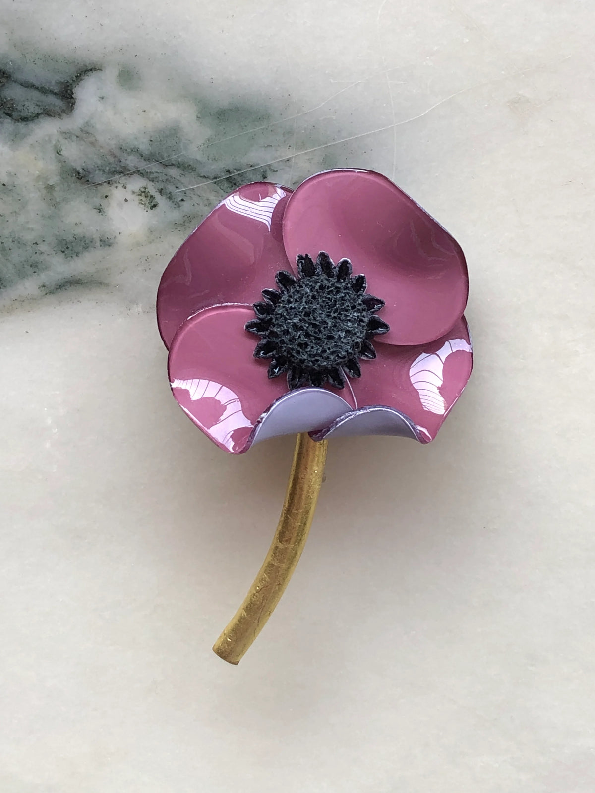 Broochella Fabrice Paris Poppy Flower Brooch in purple with glossy sculptural petals, a textured black centre and curved antique gold stem. Photographed on white marble with grey-green veining.