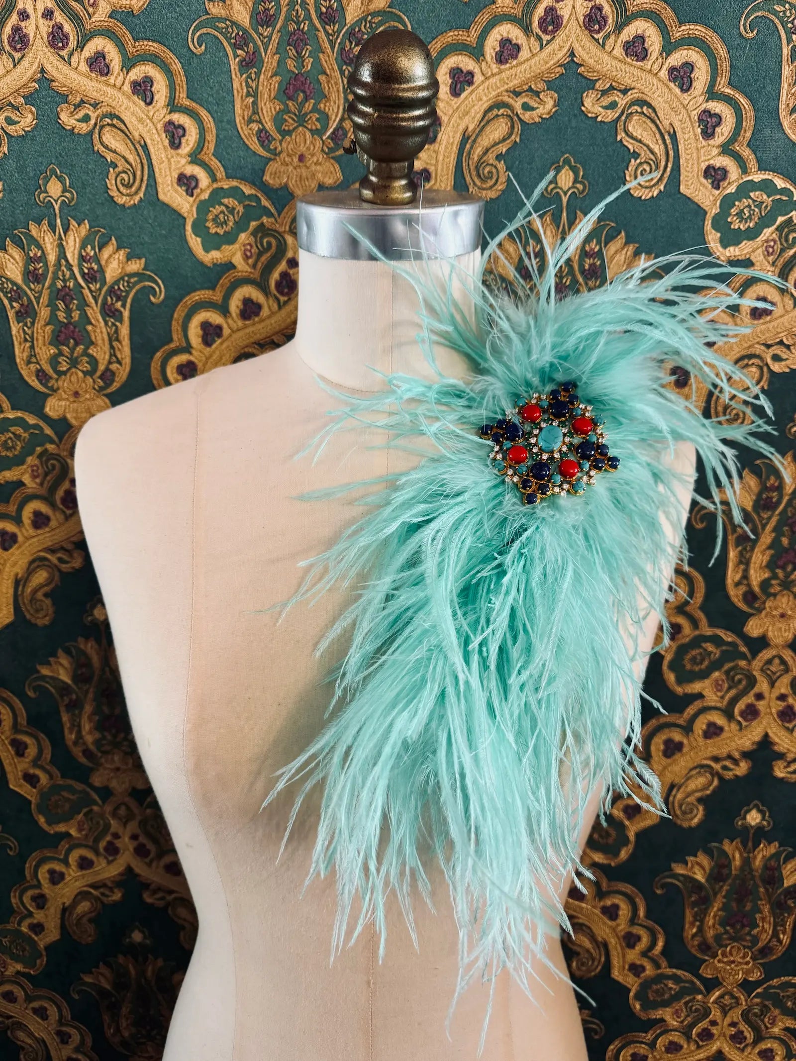 Broochella aqua handmade large feather brooch with statement brooch centre embellishment.  Pictured on a mannequin.  The background is green and gold damask wallpaper.