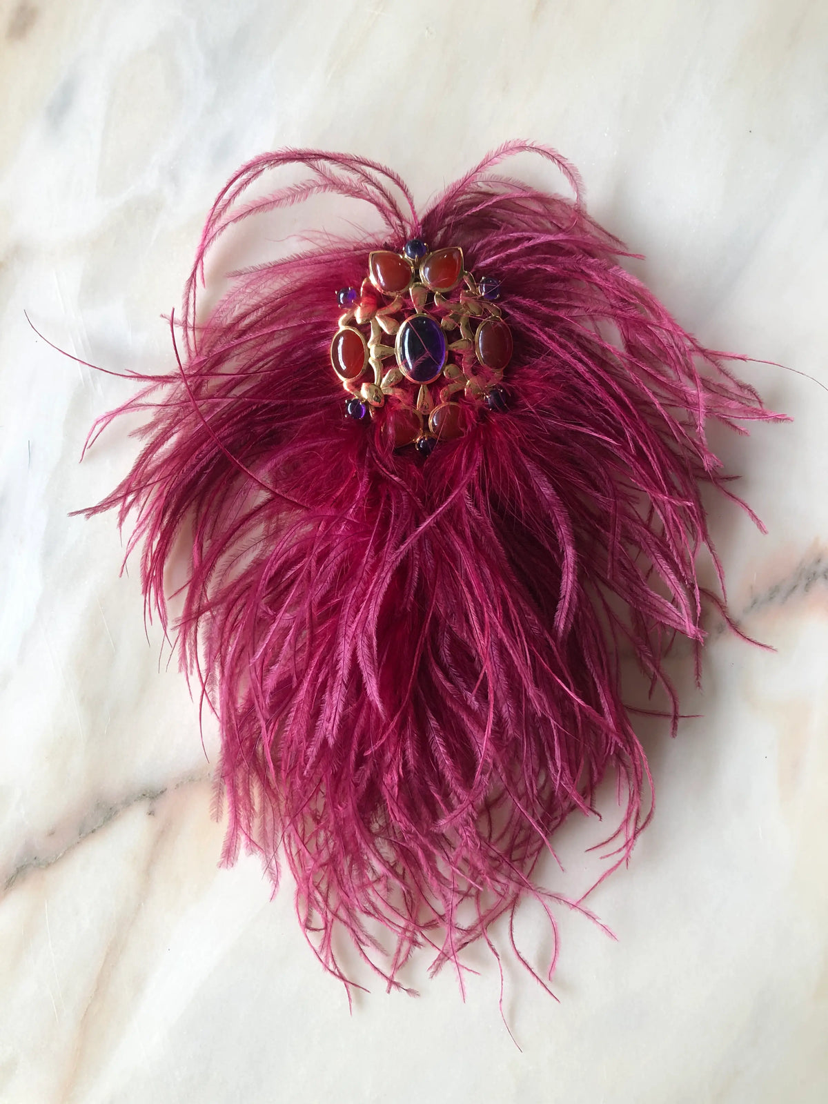 Broochella magenta Feather Bejewelled Brooch with cascading ostrich feathers and an ornate red, purple and gold-toned jewelled centre, photographed on a white marble background.