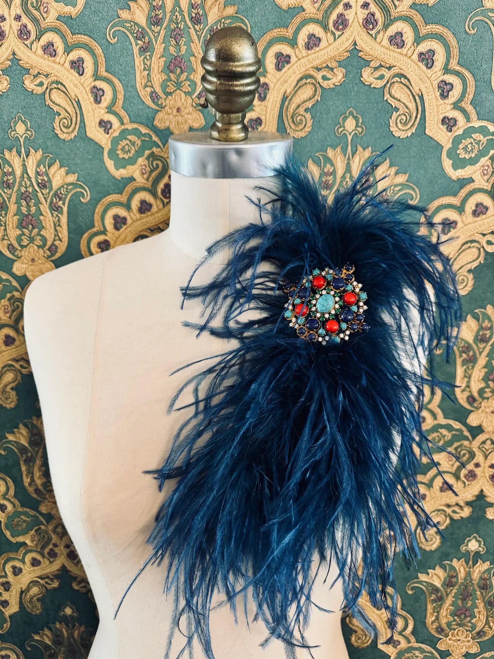 Broochella blue handmade large feather brooch with statement brooch centre embellishment.  Pictured on a mannequin.  The background is green and gold damask wallpaper.