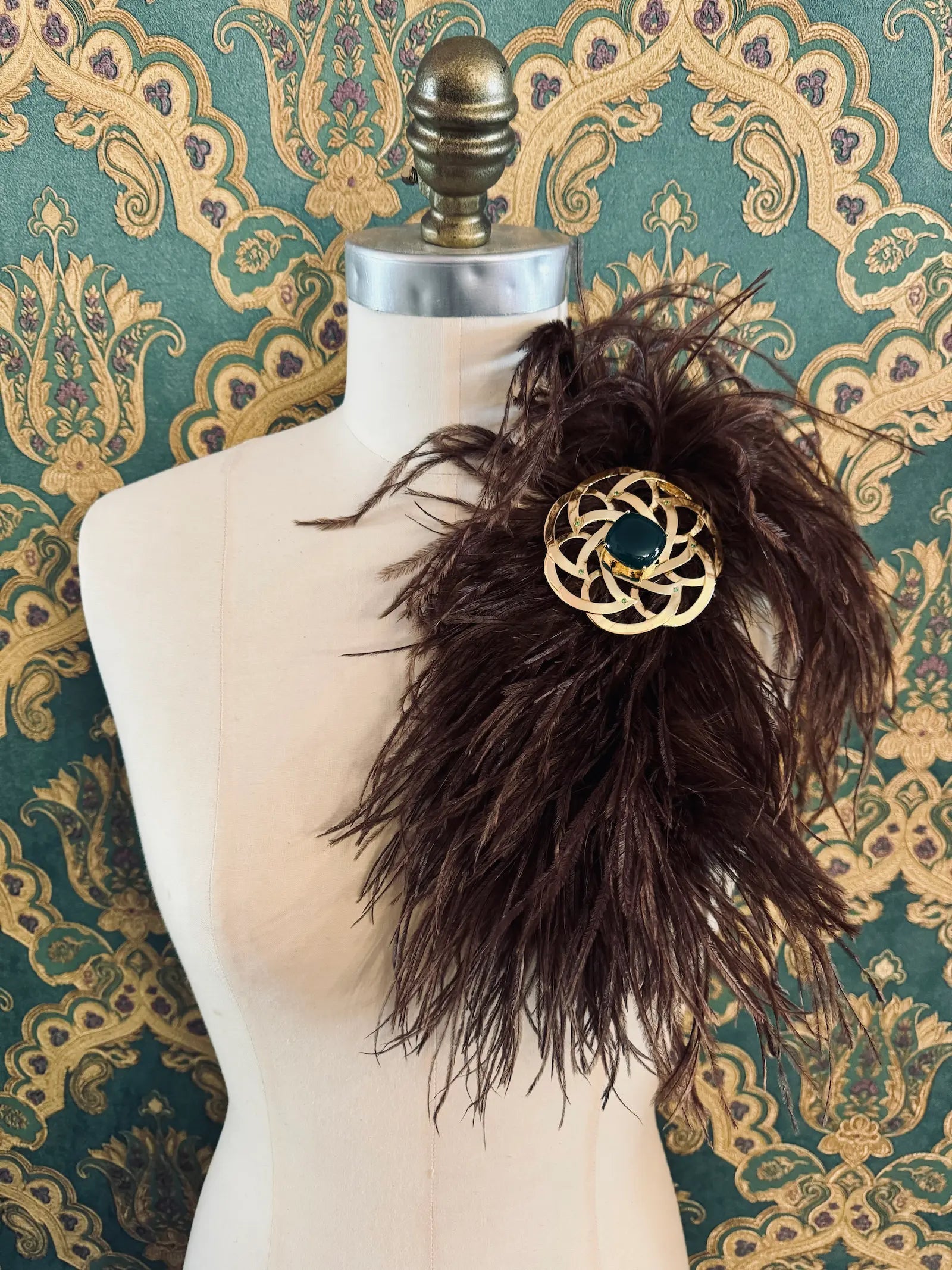 Broochella brown handmade large feather brooch with statement brooch centre embellishment.  Pictured on a mannequin.  The background is green and gold damask wallpaper.