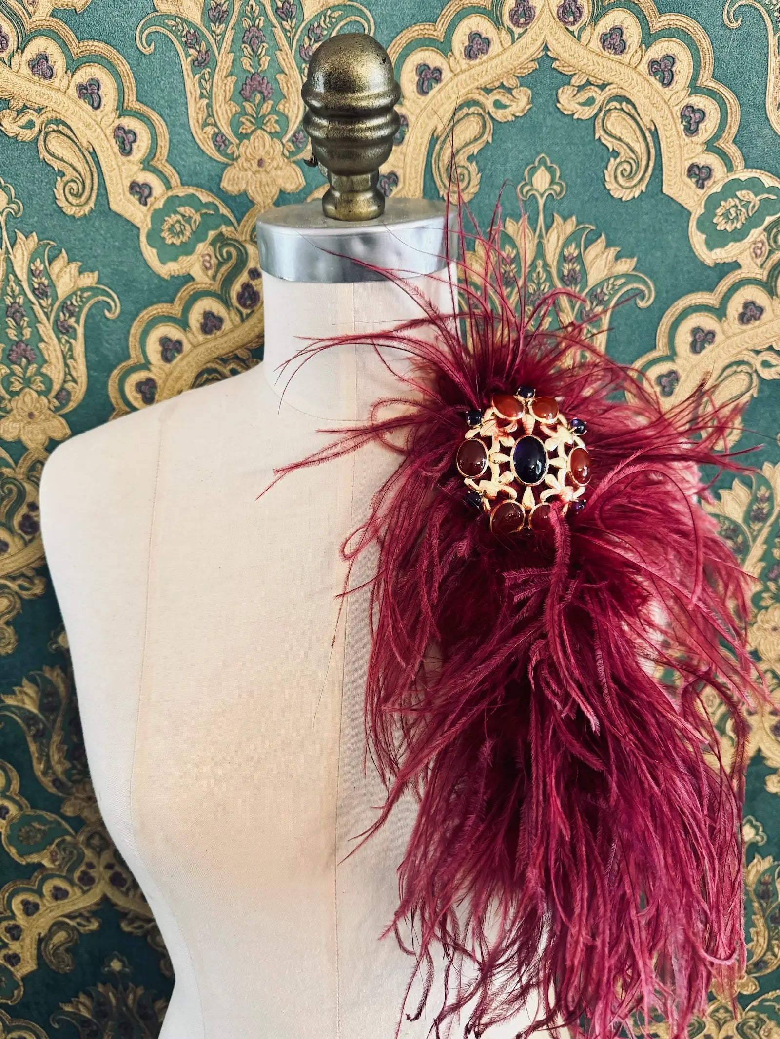 Broochella magenta handmade large feather brooch with statement brooch centre embellishment.  Pictured on a mannequin.  The background is green and gold damask wallpaper.