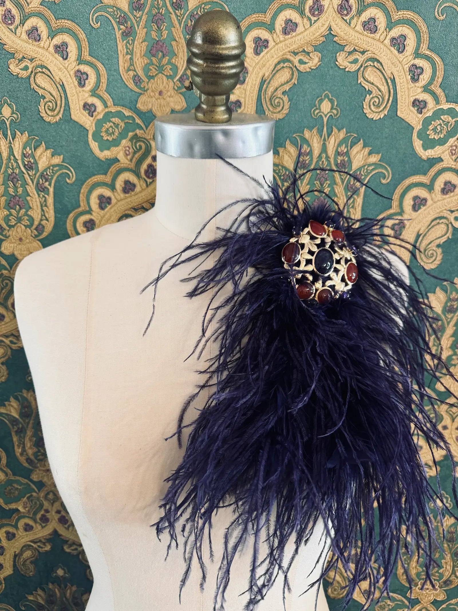 Broochella purple handmade large feather brooch with statement brooch centre embellishment.  Pictured on a mannequin.  The background is green and gold damask wallpaper.