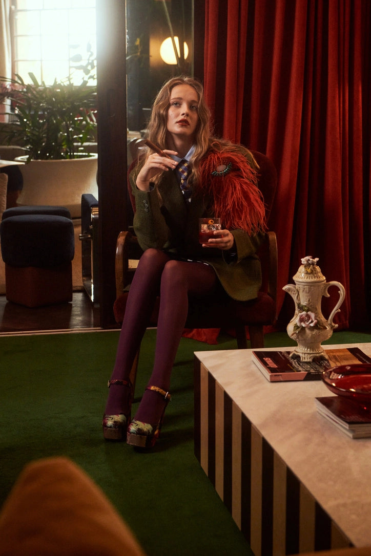 A woman sits in a chair in a stylish interior setting, wearing a dark green jacket with a  Broochella rust bejewelled feather brooch, purple tights, and purple heels. She holds a glass of water and a small glass in a dimly lit room with red curtains, a green carpet, and a striped table in the foreground.
