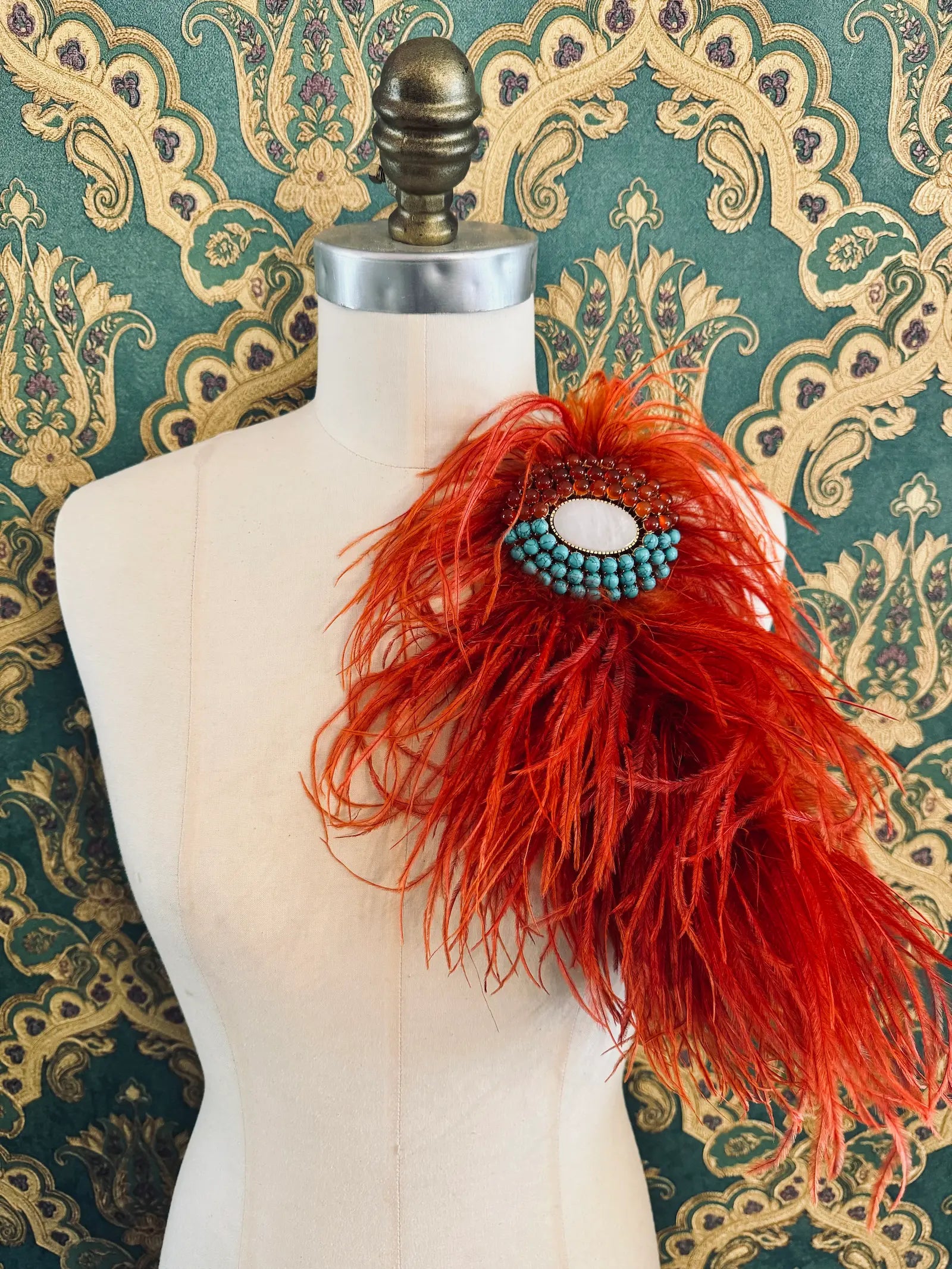 Broochella rust handmade large feather brooch with statement brooch centre embellishment.  Pictured on a mannequin.  The background is green and gold damask wallpaper.