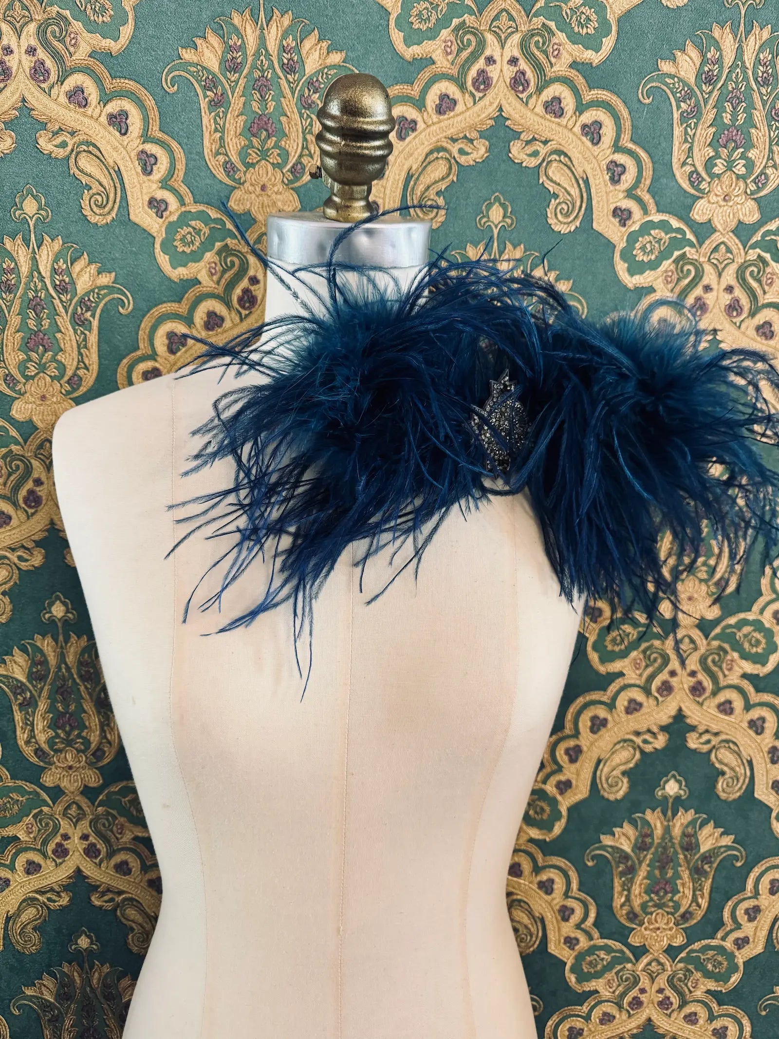 Broochella blue handmade large feather bow brooch with embellished centre.  Pictured on a mannequin.  The background is green and gold damask wallpaper.