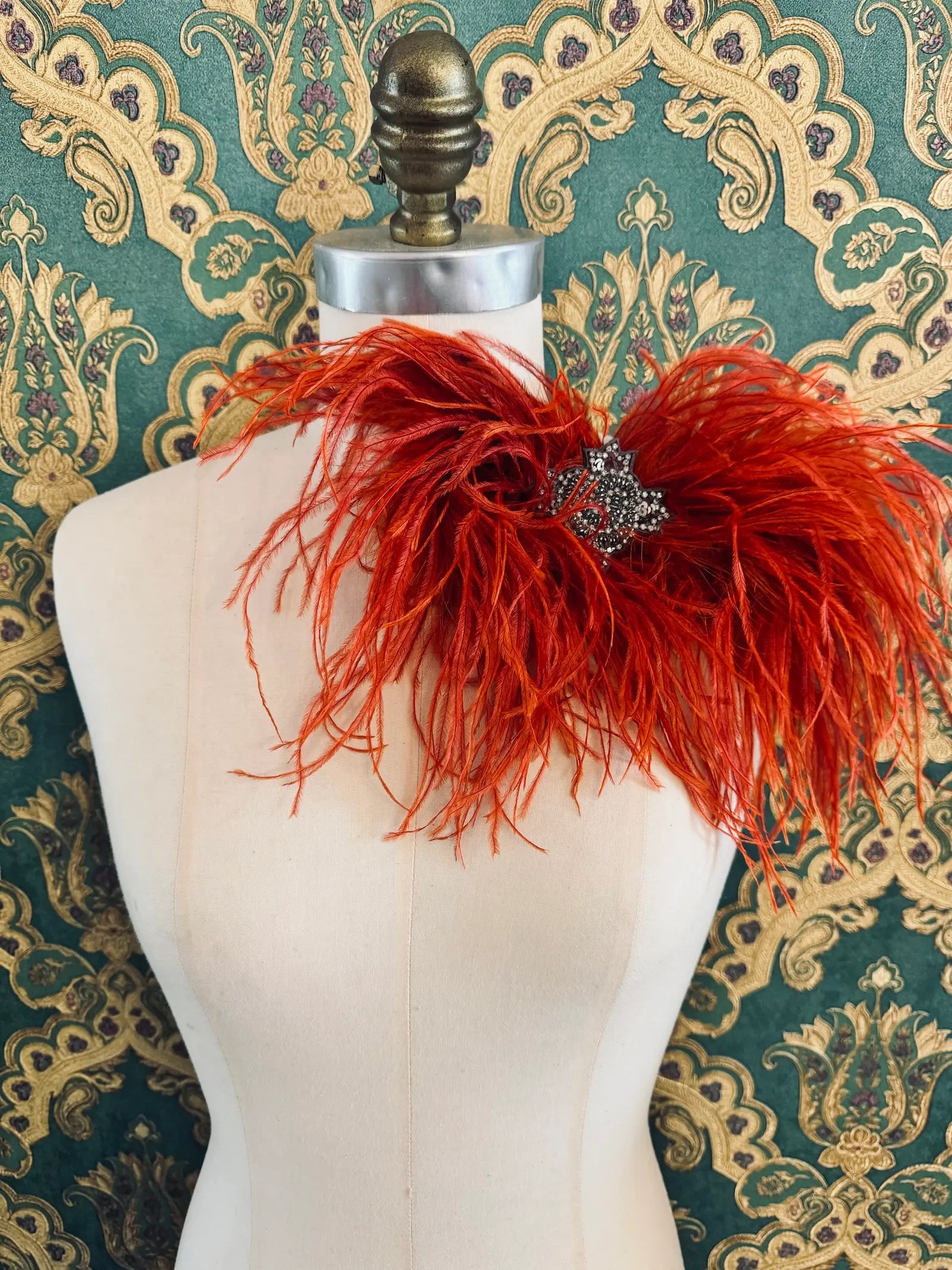 Broochella rust handmade large feather bow brooch with embellished centre.  Pictured on a mannequin.  The background is green and gold damask wallpaper.