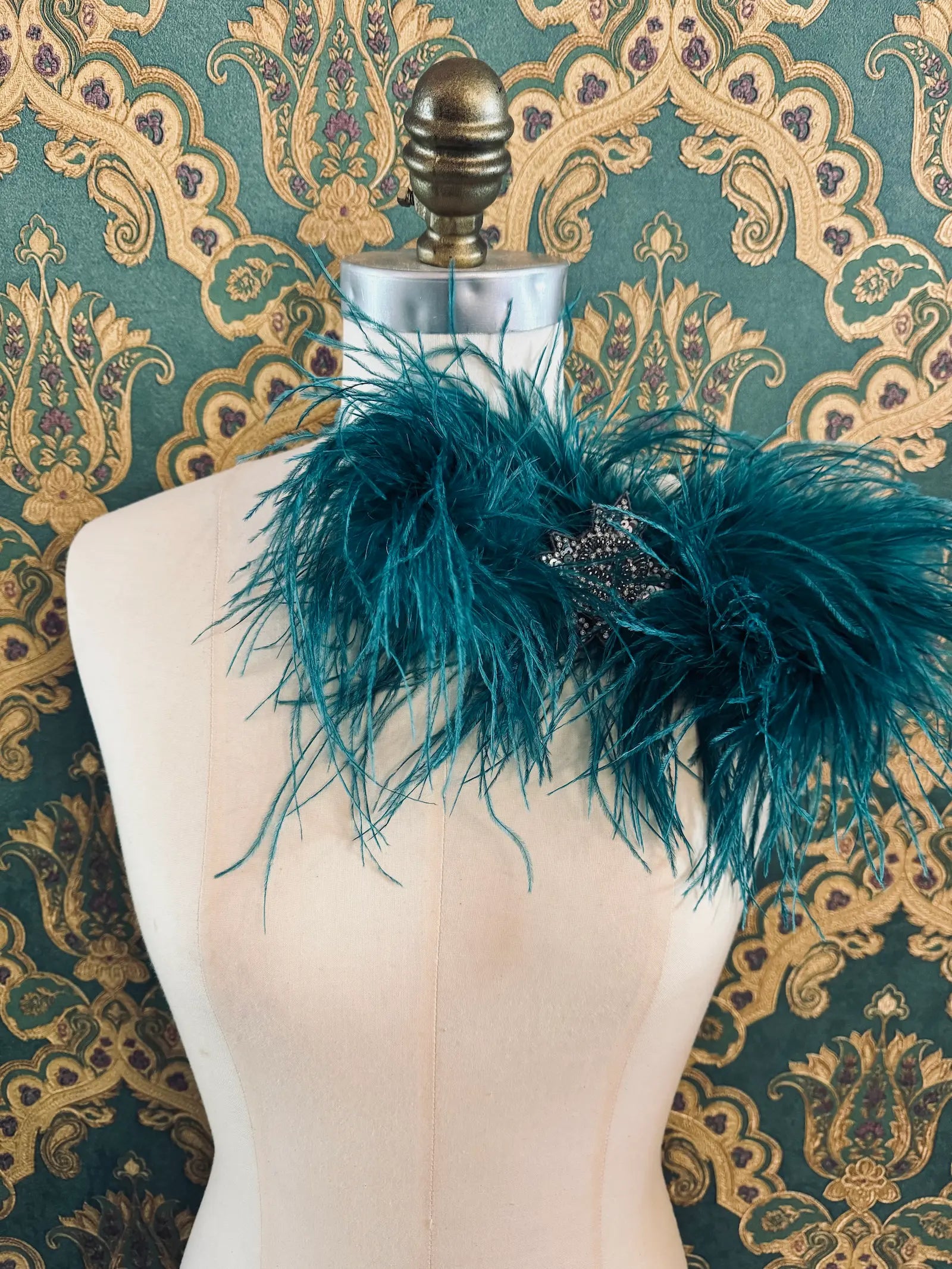 Broochella teal handmade large feather bow brooch with embellished centre.  Pictured on a mannequin.  The background is green and gold damask wallpaper.
