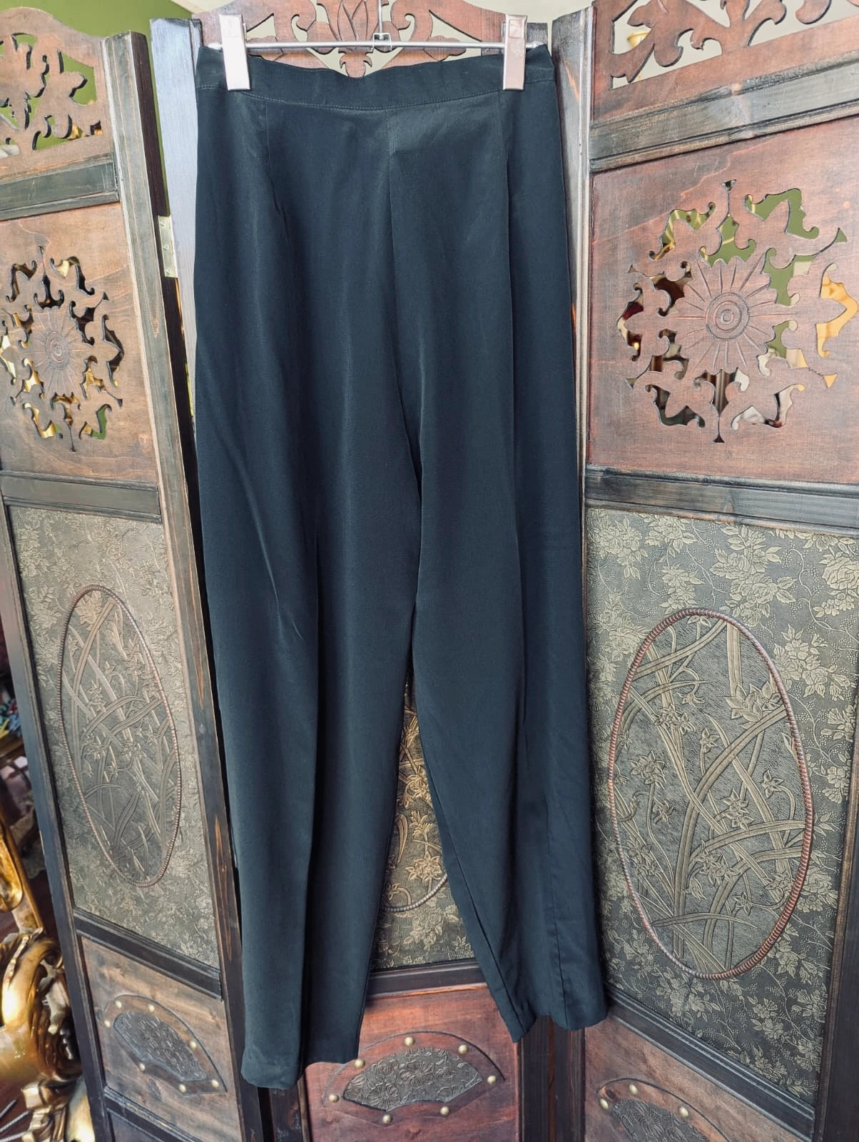 Broochella Felyne vintage black pleated front trousers. They are hung over an ortnate, dark wood room divider. 