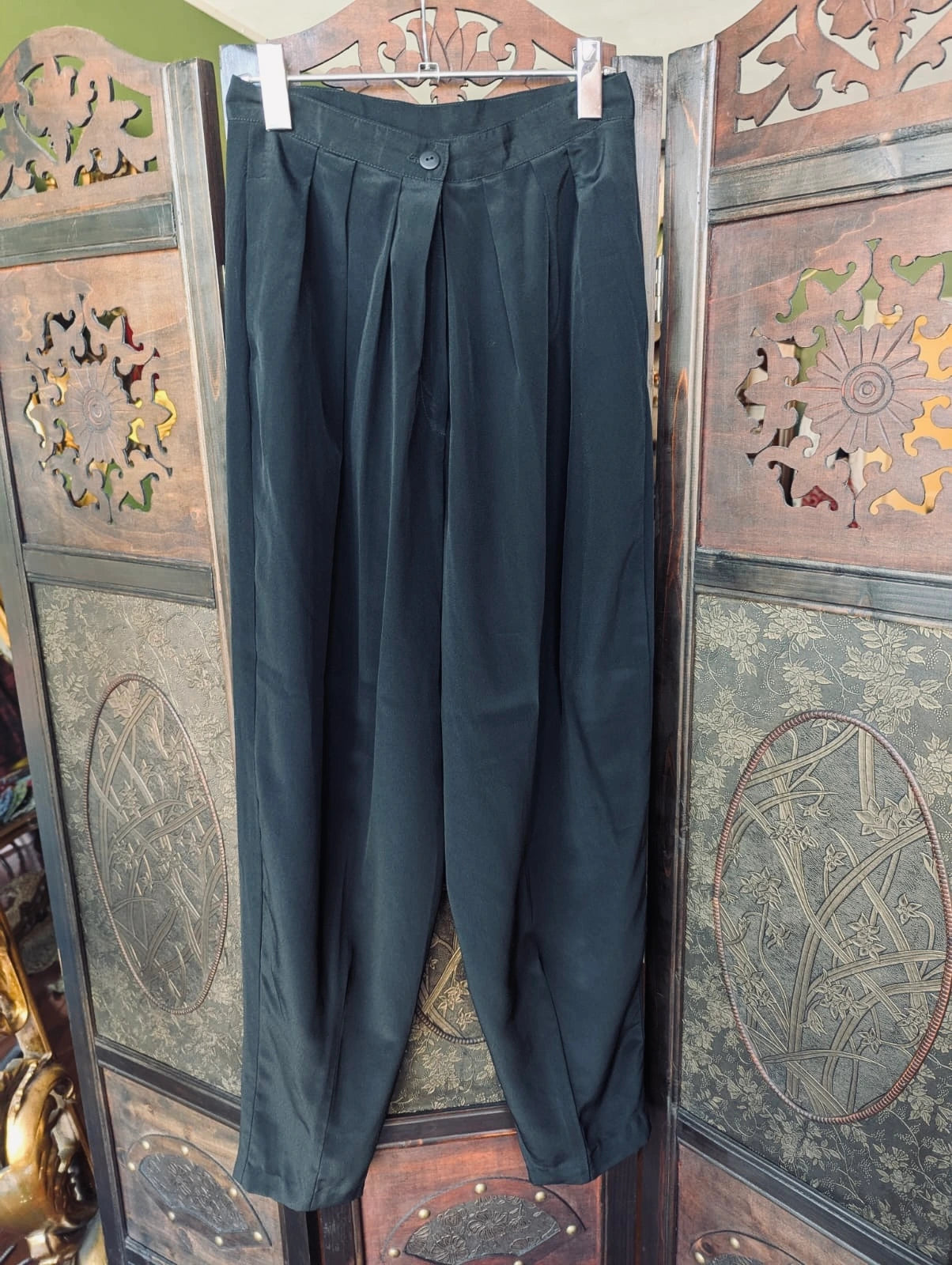 Broochella Felyne vintage black pleated front trousers. They are hung over an ortnate, dark wood room divider. 