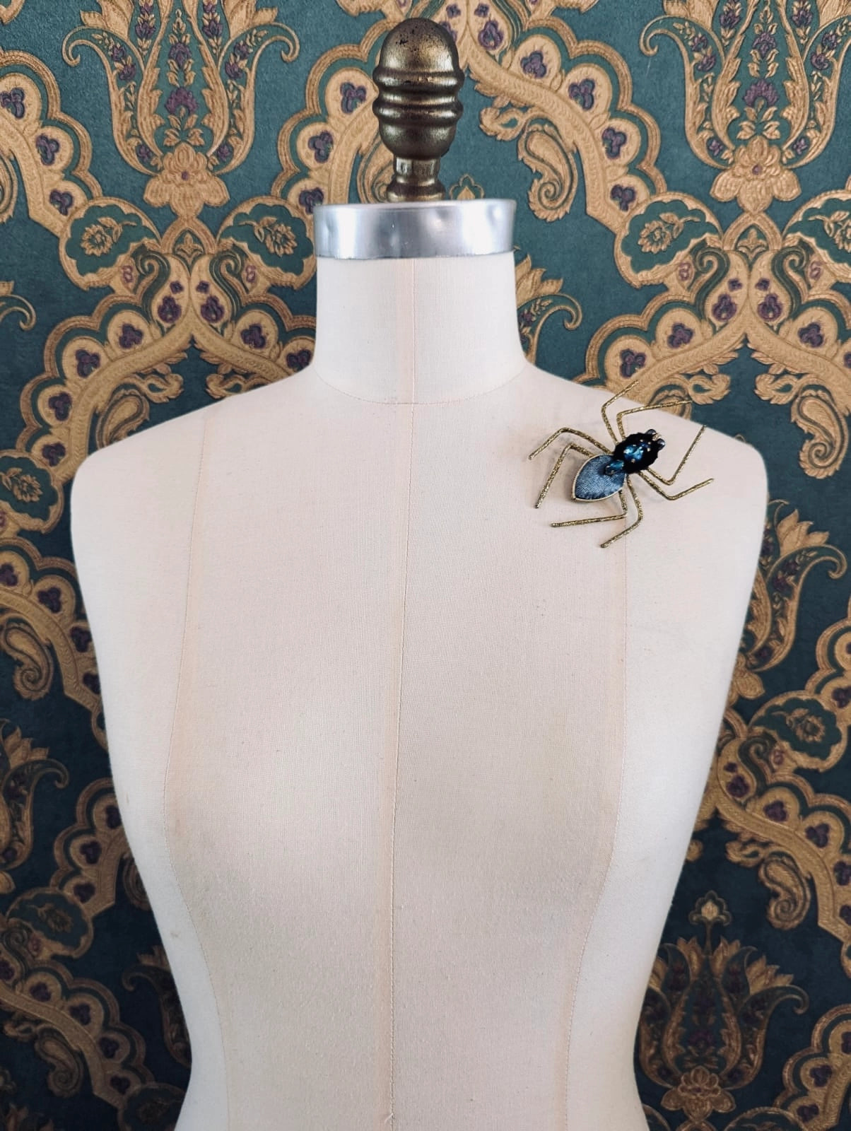 Broochella  men's viva la stella hand beaded brooch. The brooch is round, with a yellow star at the centre. There are blue, orange and white beads around the star. The brooch is pinned on a mannequin. The background is green and gold damask wallpaper.