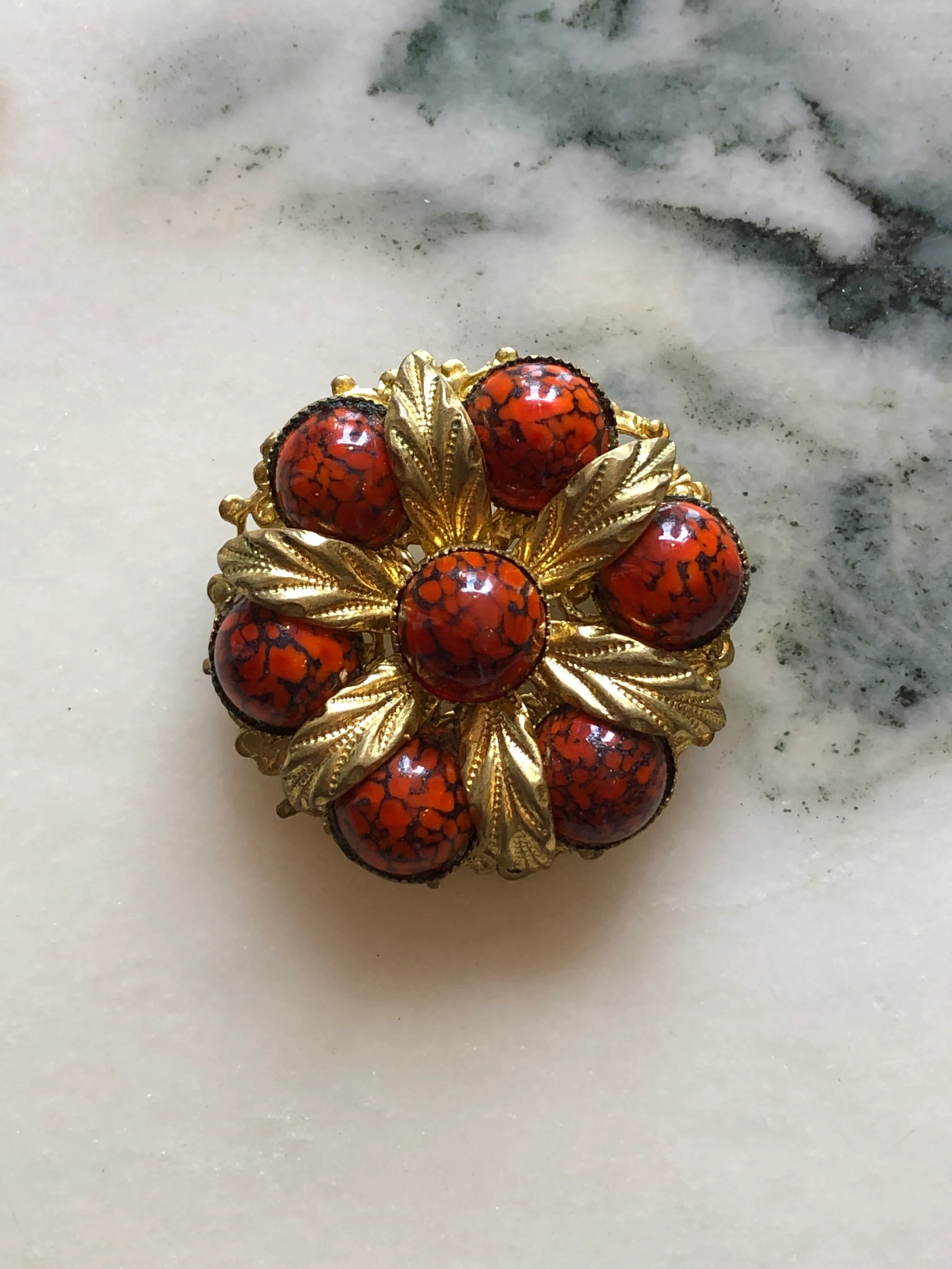 Broochella Fiamma Boema vintage Broochella Czech brooch with red-orange marbled glass cabochons and sculpted gold-tone leaves. The background is white marble.