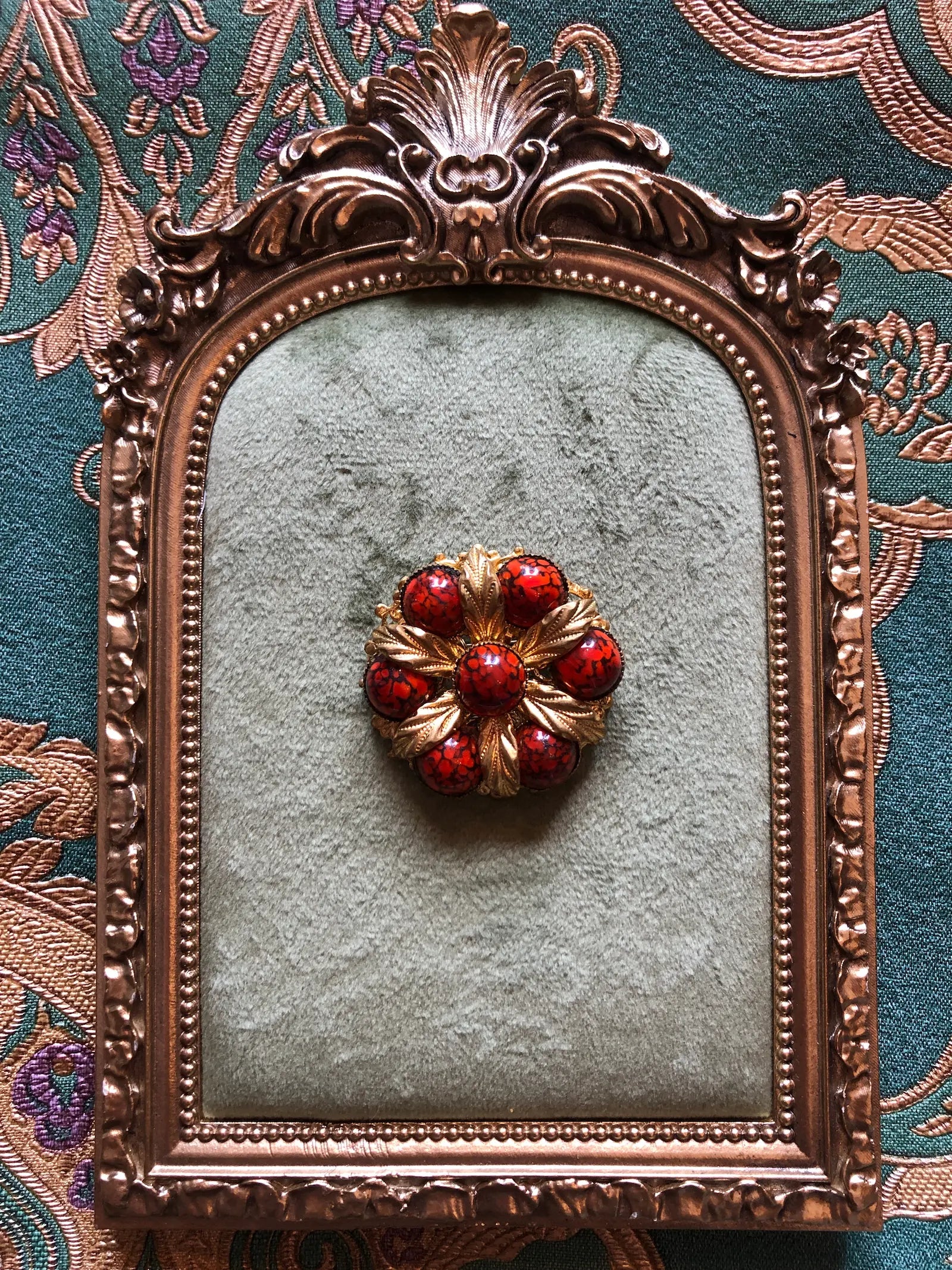 Broochella Fiamma Boema vintage Czech brooch with red-orange marbled glass cabochons and sculpted gold-tone leaves. It is displayed on a textured light green velvet insert within an ornate antique bronze frame, set against a patterned teal fabric background.