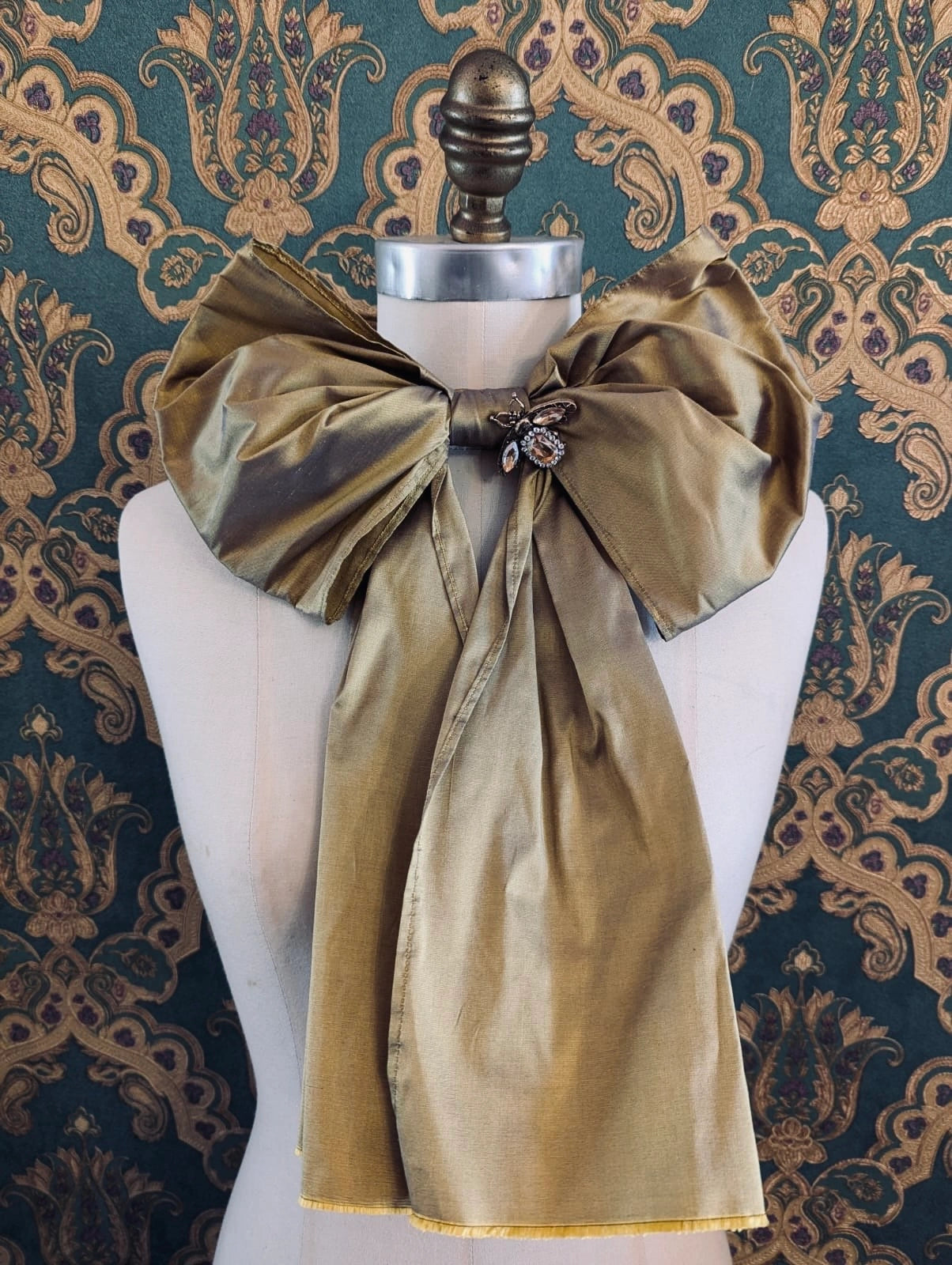 Broochella Fiocco large handmade silk embellished bow in gold. Pictured on a mannequin. The background is green and gold damask wallpaper.