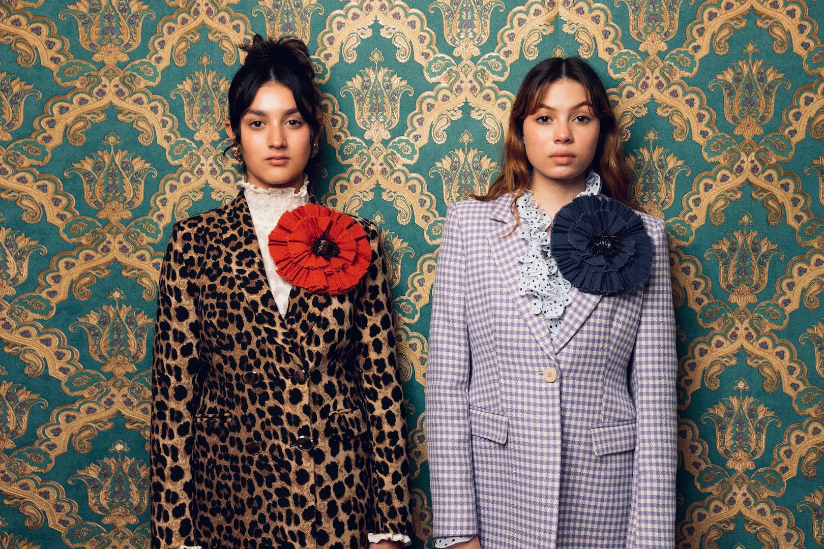 Two models standing side-by-side in front of a teal and gold damask wallpaper background. The model on the left wears a brown and black leopard print blazer with a large red Broochella fiordaliso fabric flower brooch on the lapel.