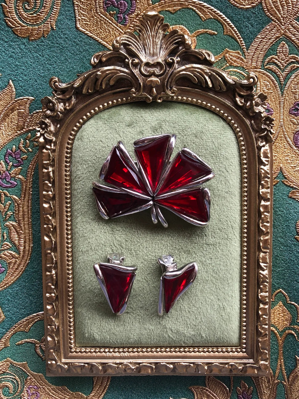 Broochella Sole d’Oro Vintage Brooch featuring sculptural gold-tone leaves and an iridescent rainbow-effect centre, displayed on sage green velvet in an ornate antique-style gold frame against green and gold patterned fabric.