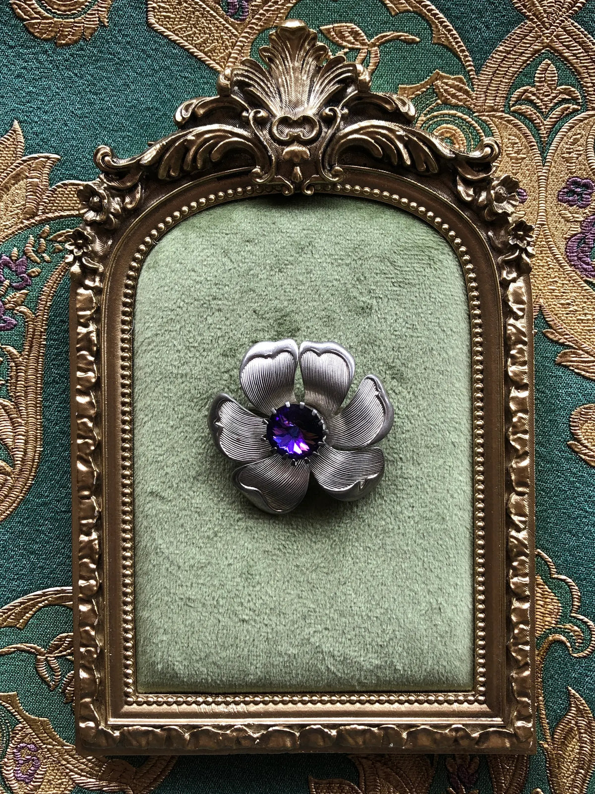 Broochella Fiore Viola Vintage Brooch, a sculptural silver-tone flower brooch with finely textured petals and a faceted violet purple crystal centre, displayed on sage green velvet in an ornate antique-style frame.