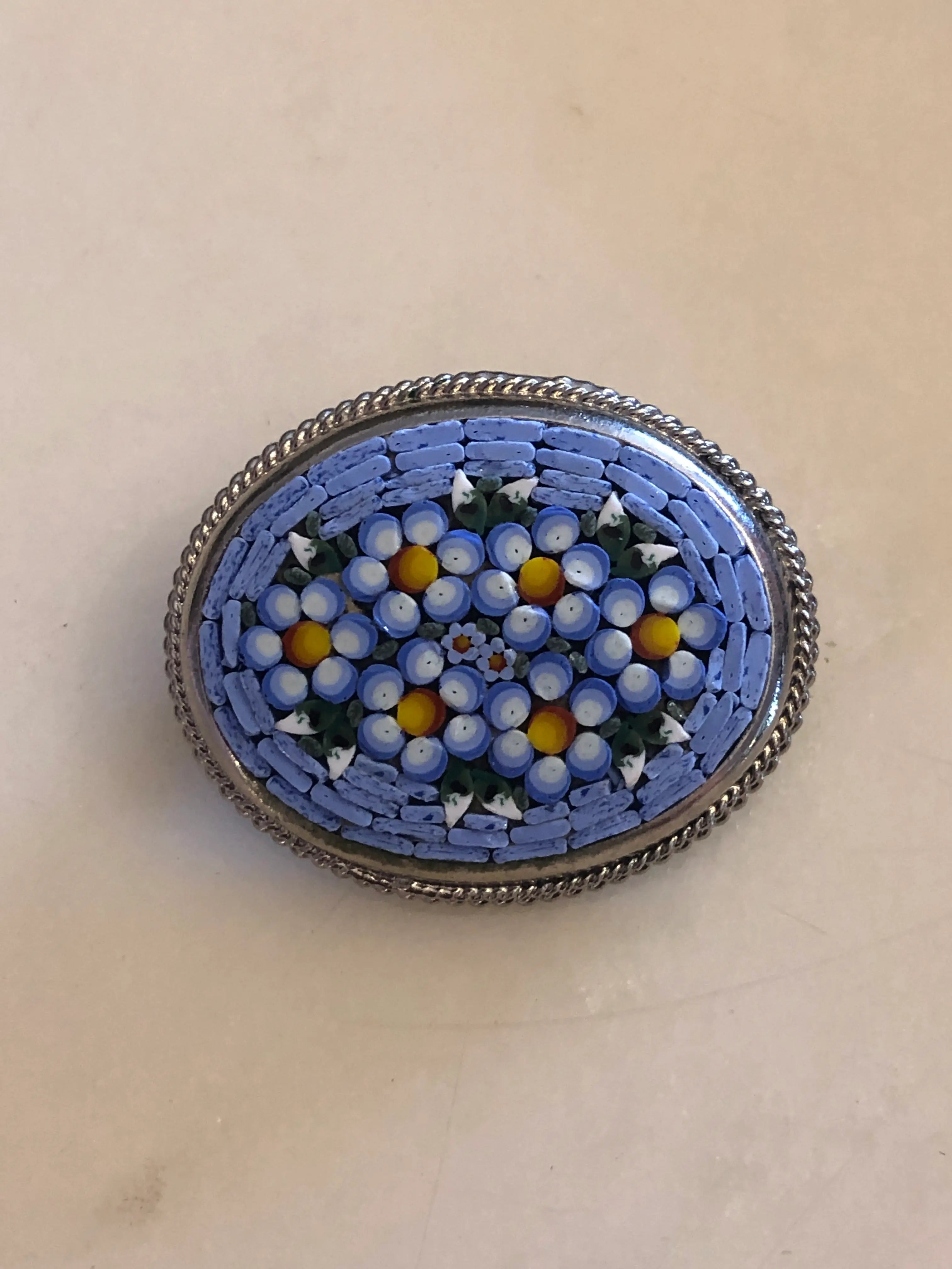 A 3cm x 4cm vintage Italian micro mosaic oval brooch featuring a hand assembled glass tessera design of forget-me-not flowers in cornflower blue, white and yellow on a blue ground, set within a silver-tone frame with twisted rope edge detail, photographed on a white marble table