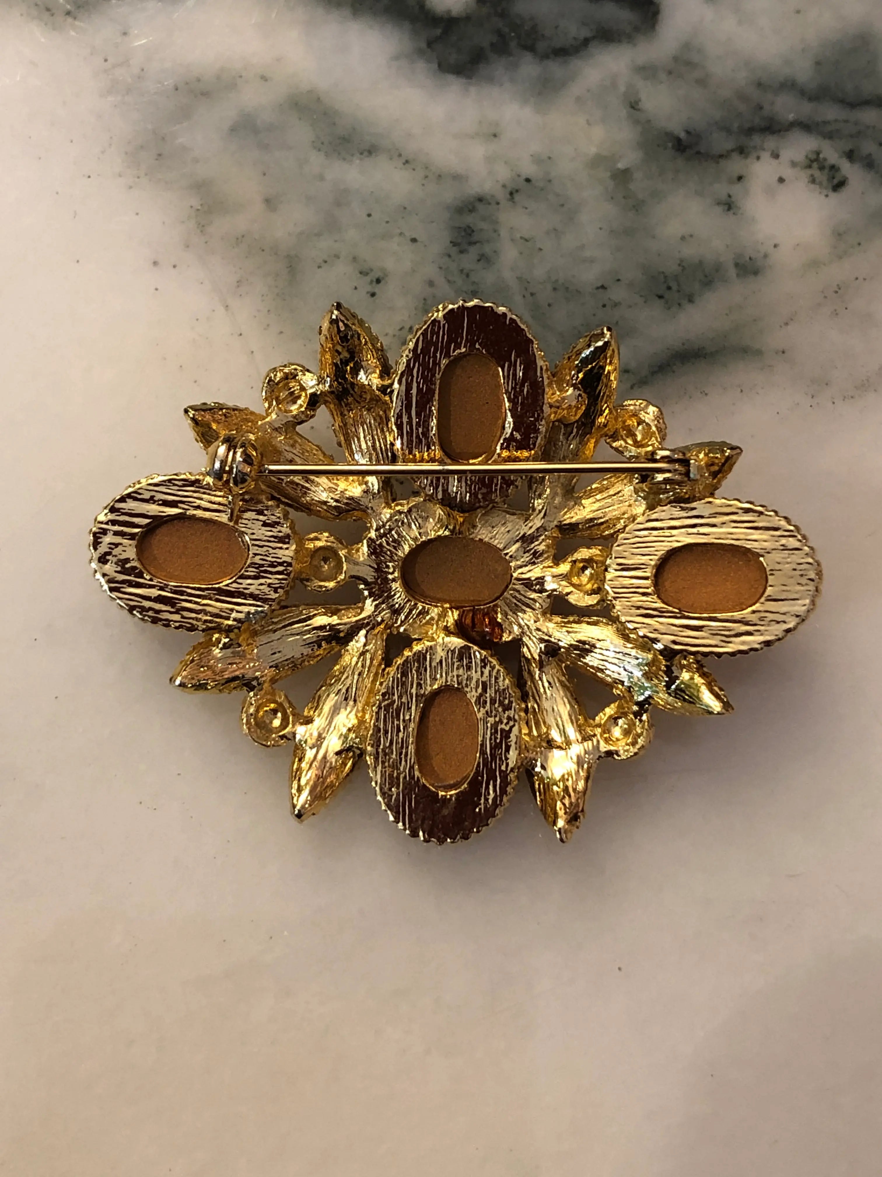 The back view of Broochella Fiorella Vintage Ruby Cabochon Brooch featuring five red oval cabochons surrounded by textured gold-tone leaves and scrollwork on a cream and grey marble background.
