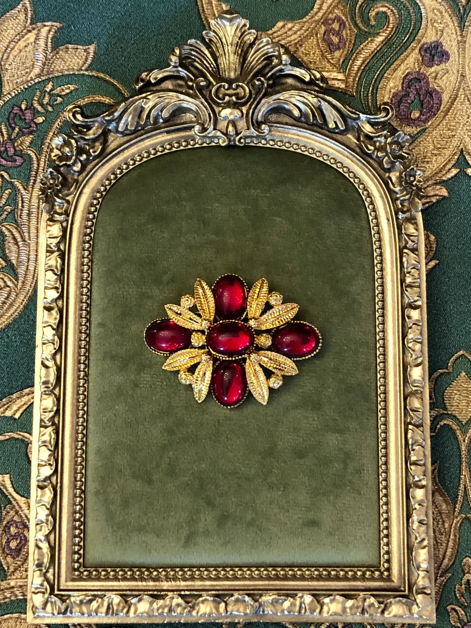 Broochella Fiorella Vintage Ruby Cabochon Brooch featuring five red oval cabochons surrounded by textured gold-tone leaves and scrollwork. Pictured on a green velvet tray with an ornate gold frame. The background is green and gold damask wallpaper