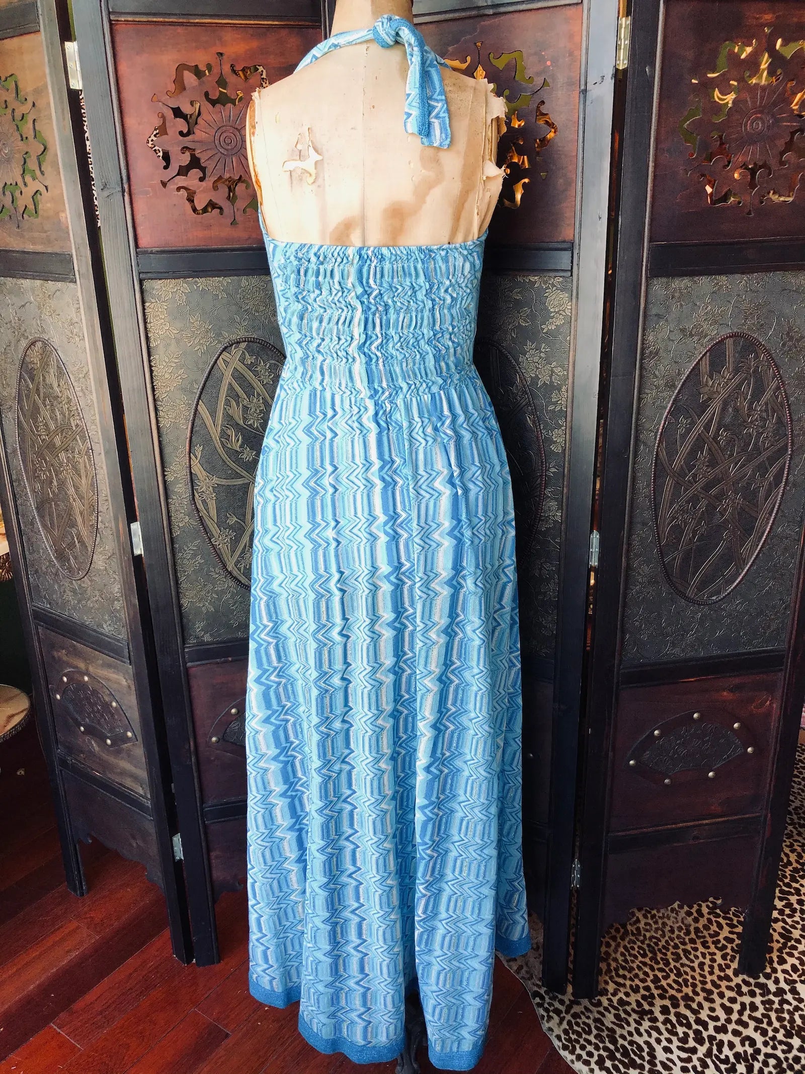 Broochella Pho Firenze maxi dress back view.  Size medium. It is a blue, white and silver zigzag pattern and is made from  46% Cotton, 46% Polyester, 8% Lurex. It has a 
halter neckline, is maxi length and has a lightweight, flowing silhouette. It is pictured on a vintage mannequin The background is an ornate, dark wood, vintage room divider. 