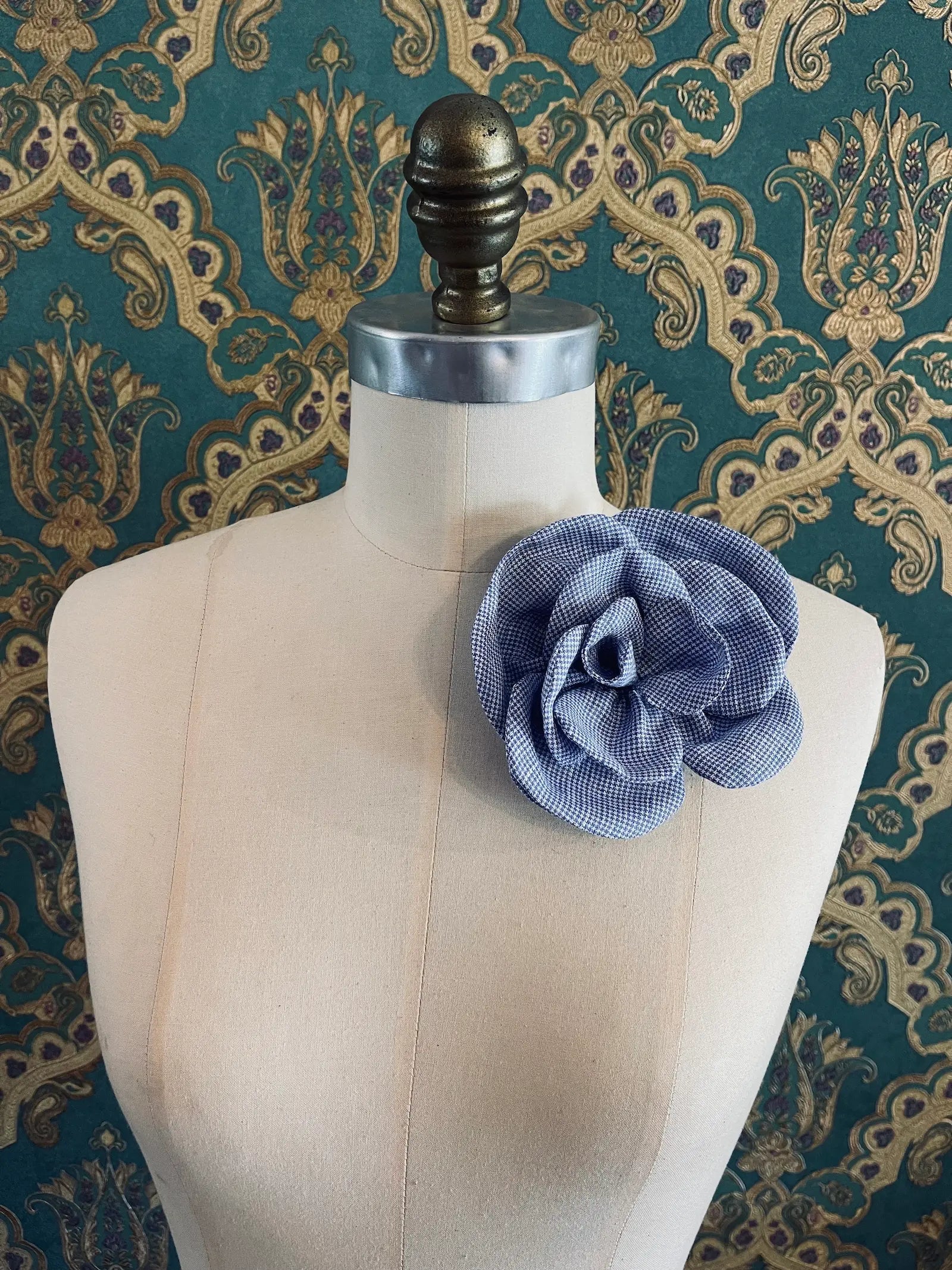 Broochella Florella blue handmade checked linen flower brooch. Pictured on a mannequin. The background is green and gold damask wallpaper