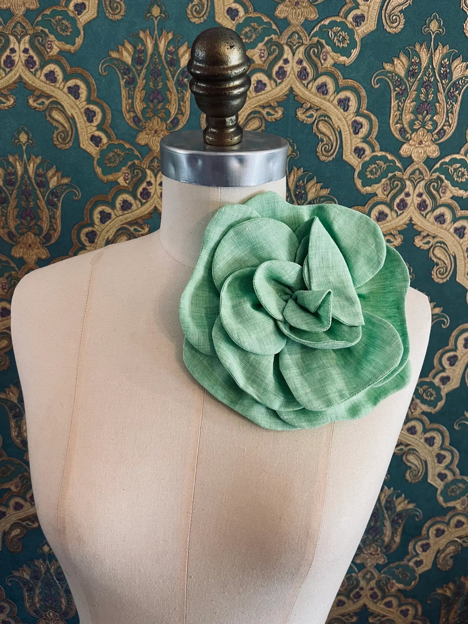 Broochella Florella large handmade linen flower brooch in green. Pictured on a mannequin. The background is green and gold damask wallpaper.