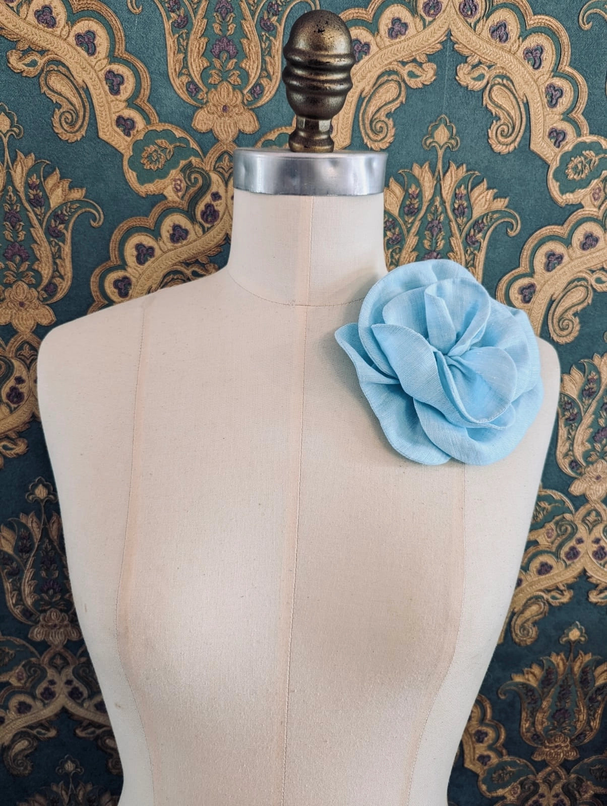 Broochella Florella small handmade linen flower brooch in blue. Pictured on a mannequin. The background is green and gold damask wallpaper.