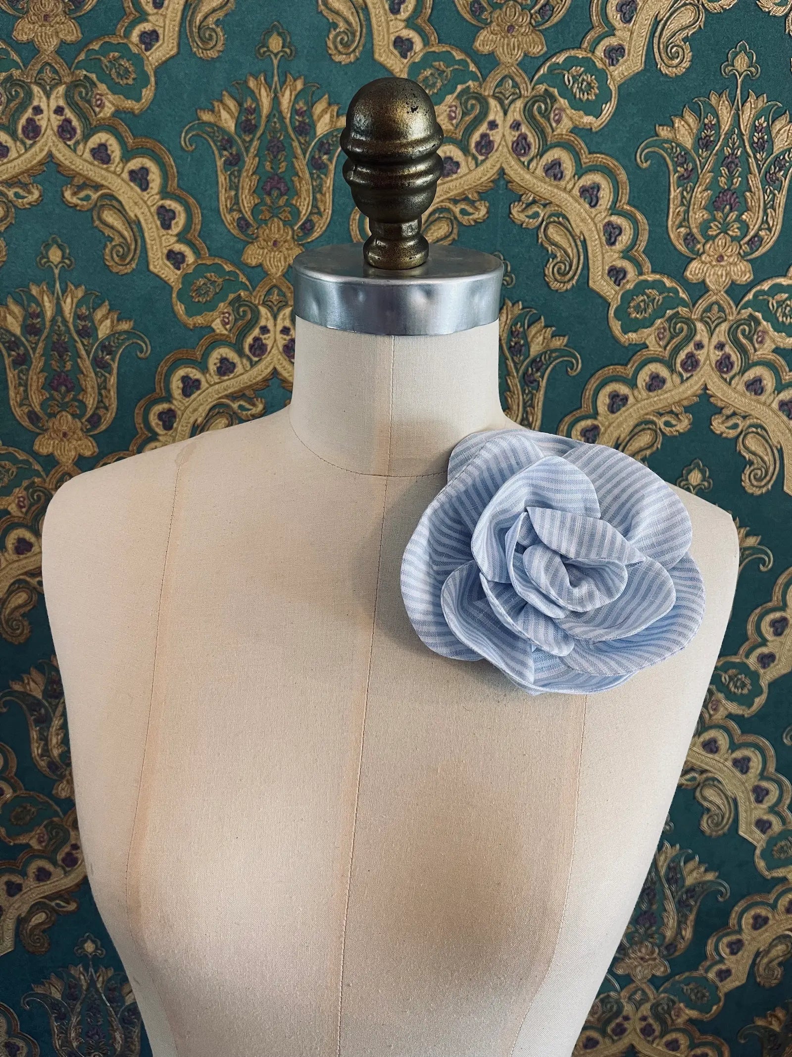 Broochella Florella blue handmade striped linen flower brooch. Pictured on a mannequin. The background is green and gold damask wallpaper