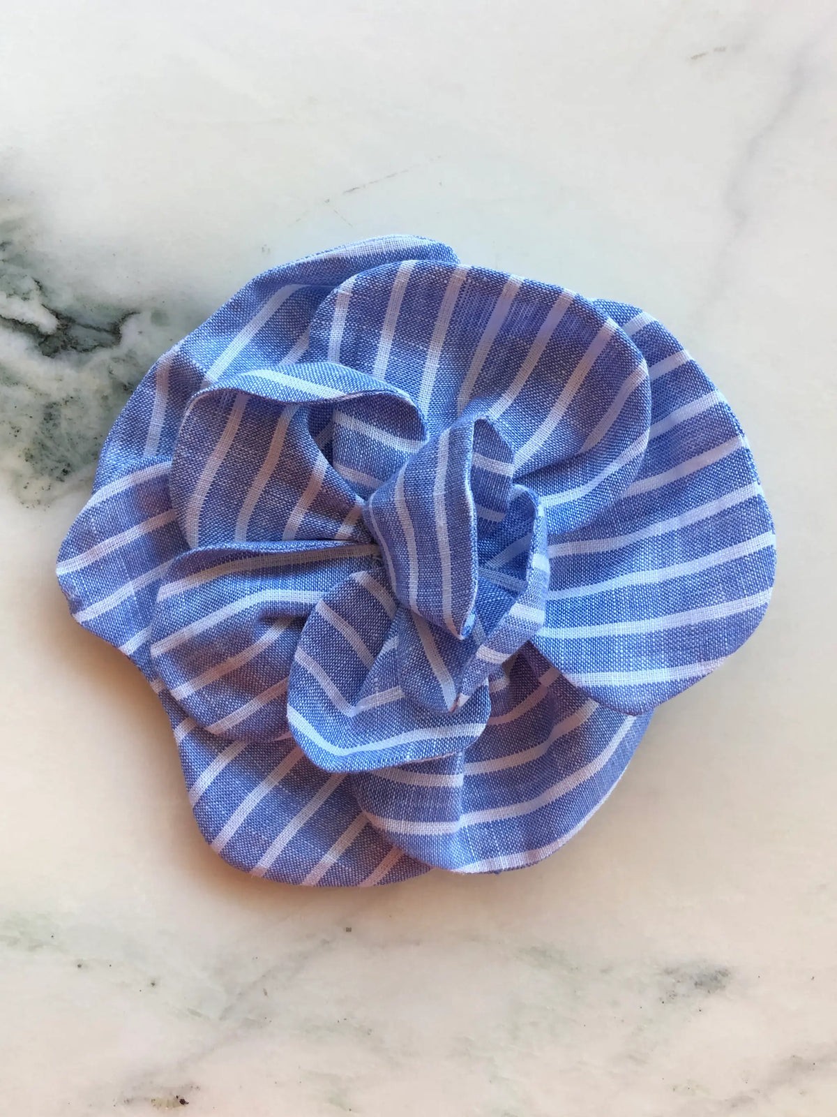 Broochella Florella blue and white striped linen flower brooch with sculptural layered petals, photographed on a white marble background with grey veining.