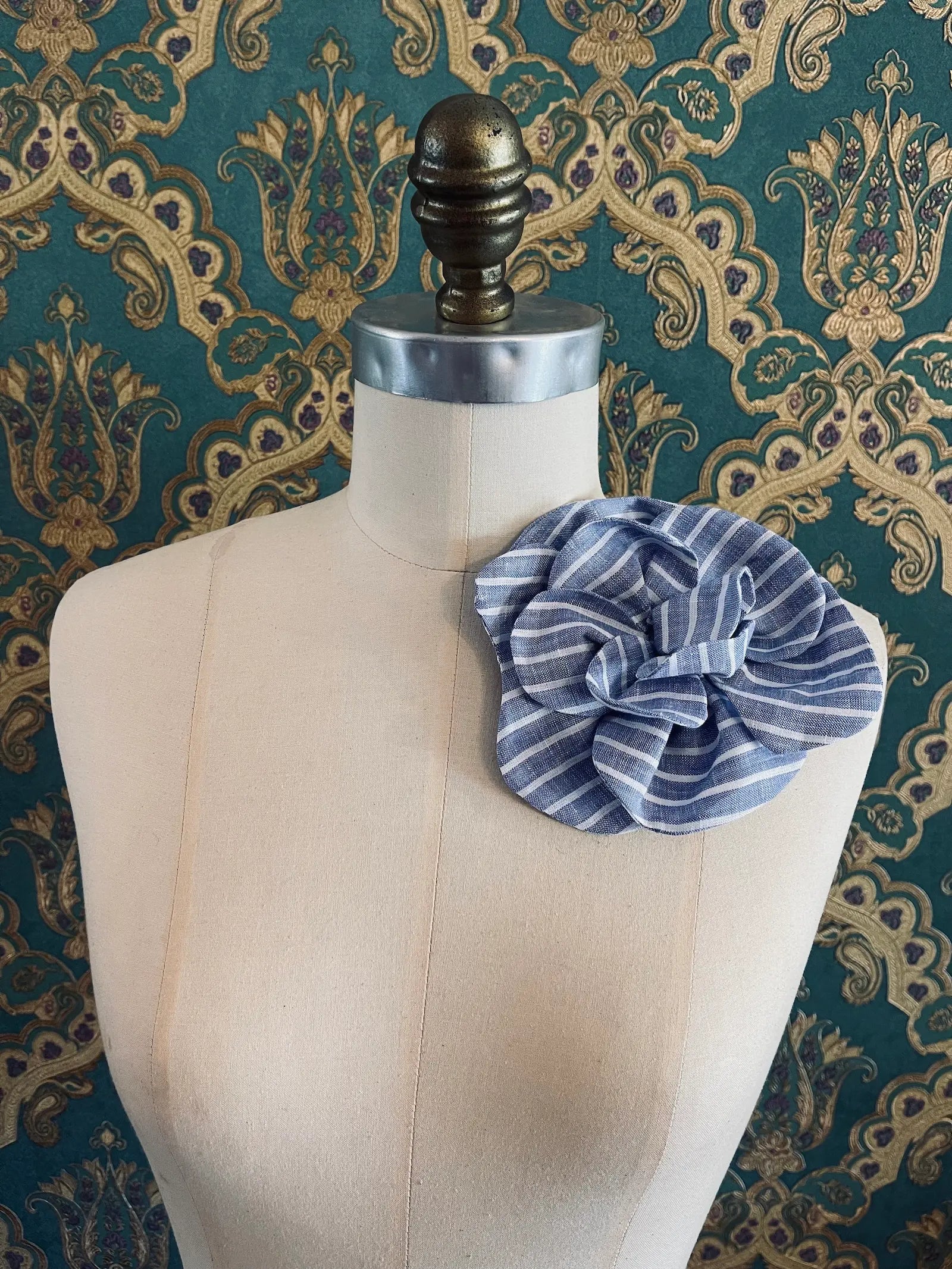 Broochella Florella blue handmade striped linen flower brooch. Pictured on a mannequin. The background is green and gold damask wallpaper