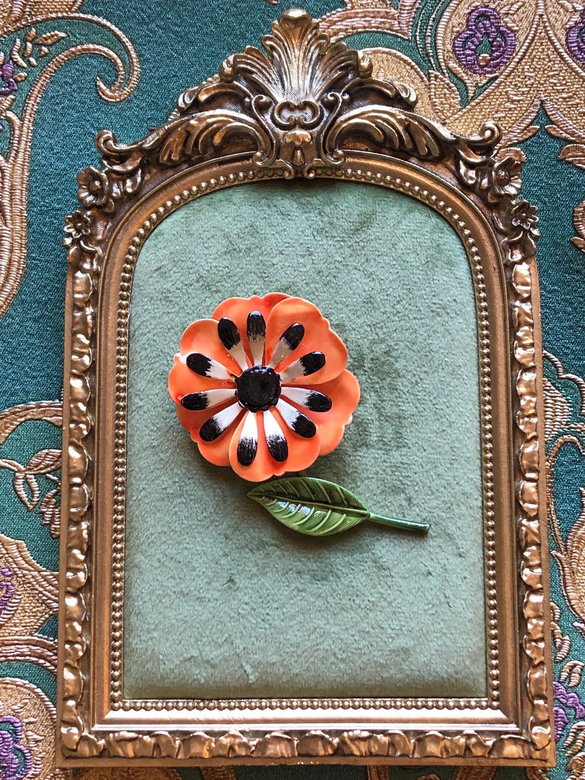 Broochella Francesca Vintage Orange Enamel Flower Brooch featuring layered orange enamel petals, black and white detailing and a green leaf and stem. Pictured on a green velvet tray with an ornate gold frame. The background is green and gold damask wallpaper
