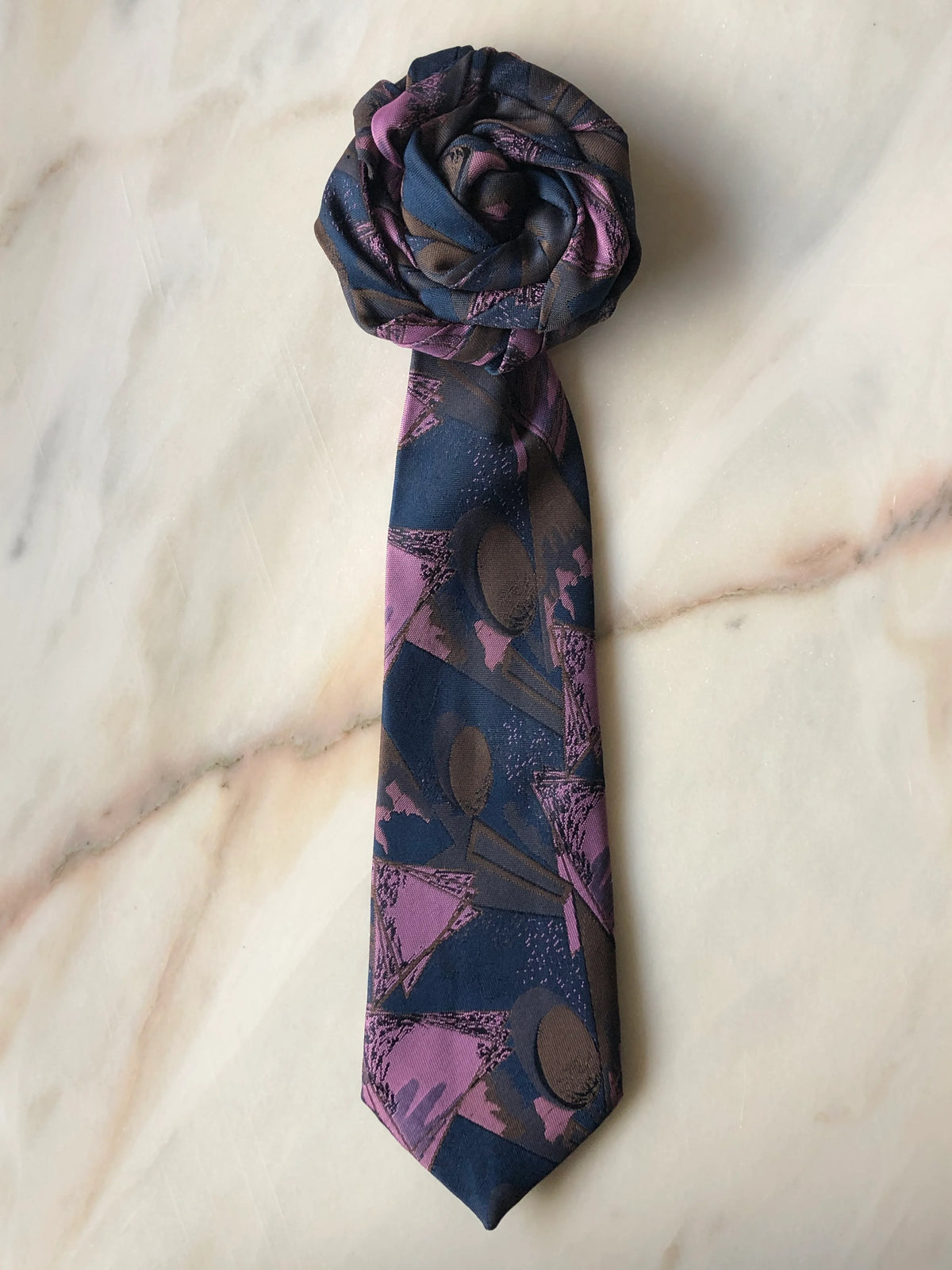 Broochella Fuente Rosette Tie Brooch - Carmelo in navy, mauve and taupe patterned fabric, finished with a sculptural rosette and photographed on a white marble background.