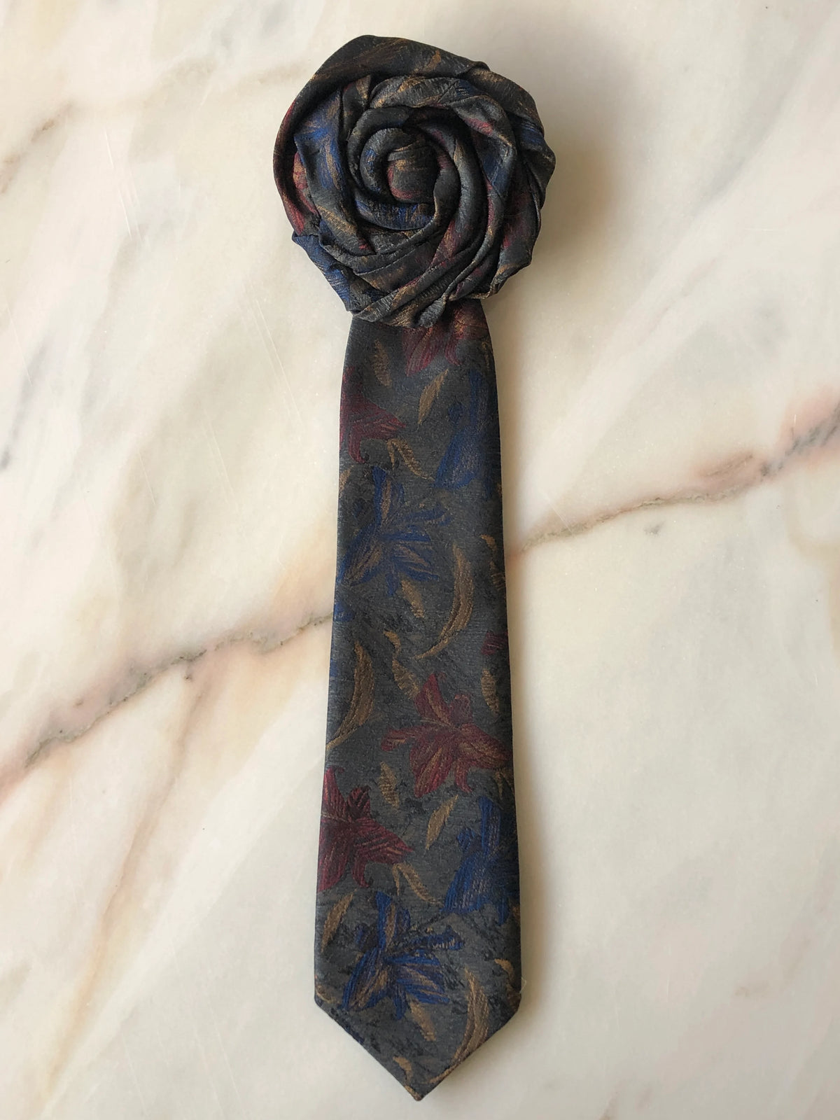 Broochella Fuente Rosette Tie Brooch - Ignazio in charcoal grey with deep blue, burgundy and bronze floral detailing, finished with a sculptural rosette and photographed on a white marble background.